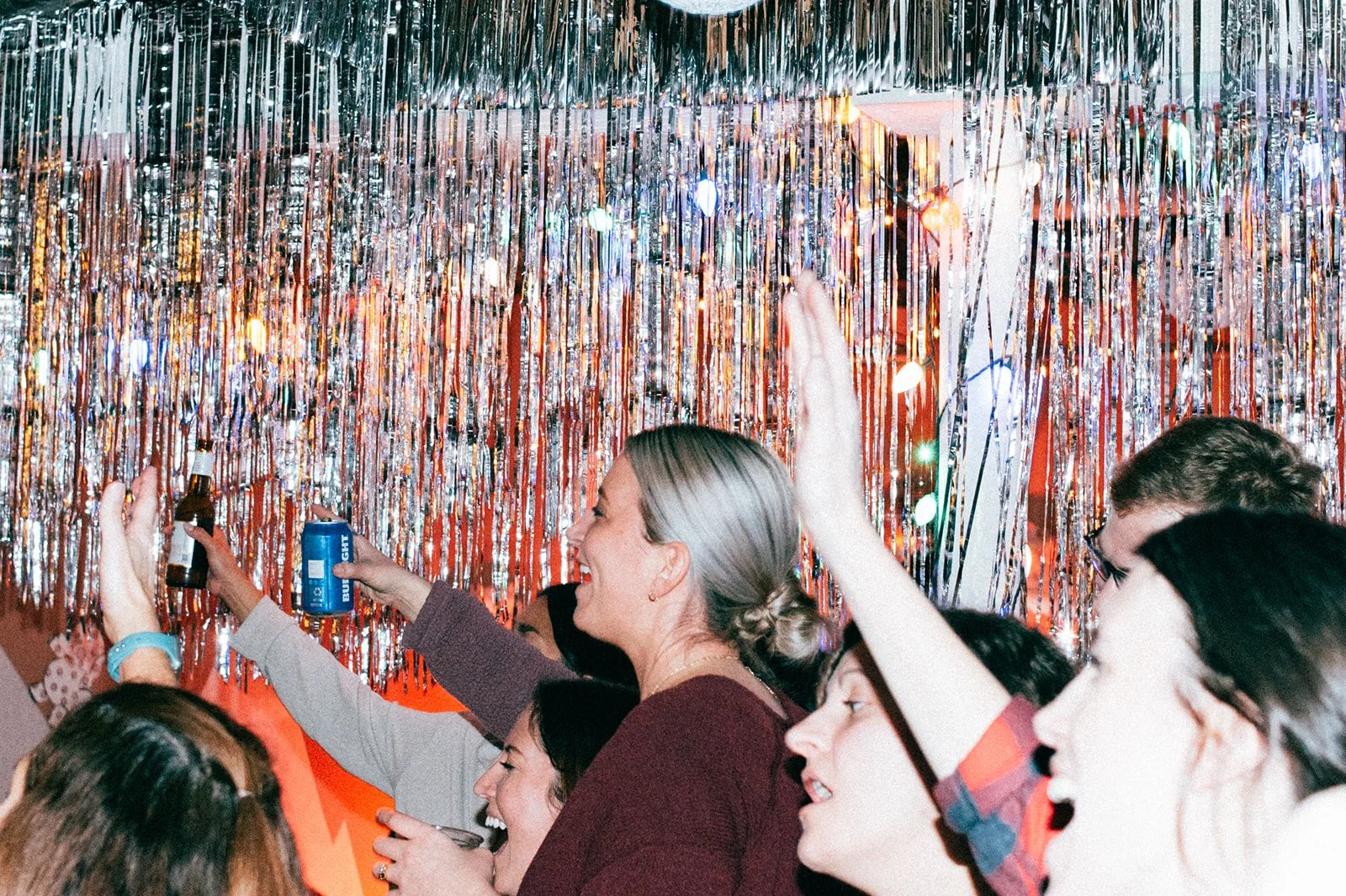 People celebrating at a party with metallic streamers and colorful lights, holding drinks and socializing.