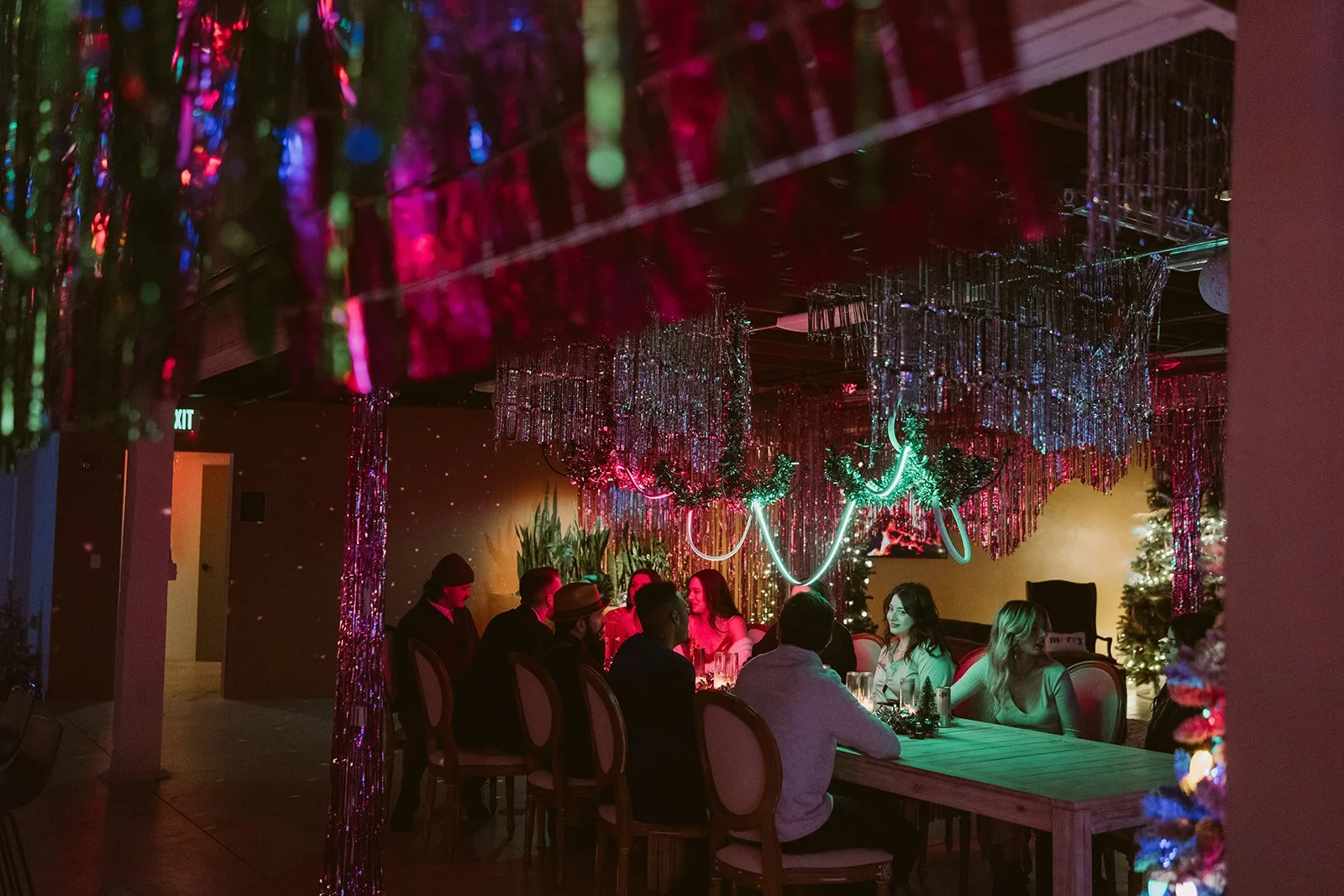 People gathered around a table at a festive holiday party with Christmas trees, tinsel, and neon lights.