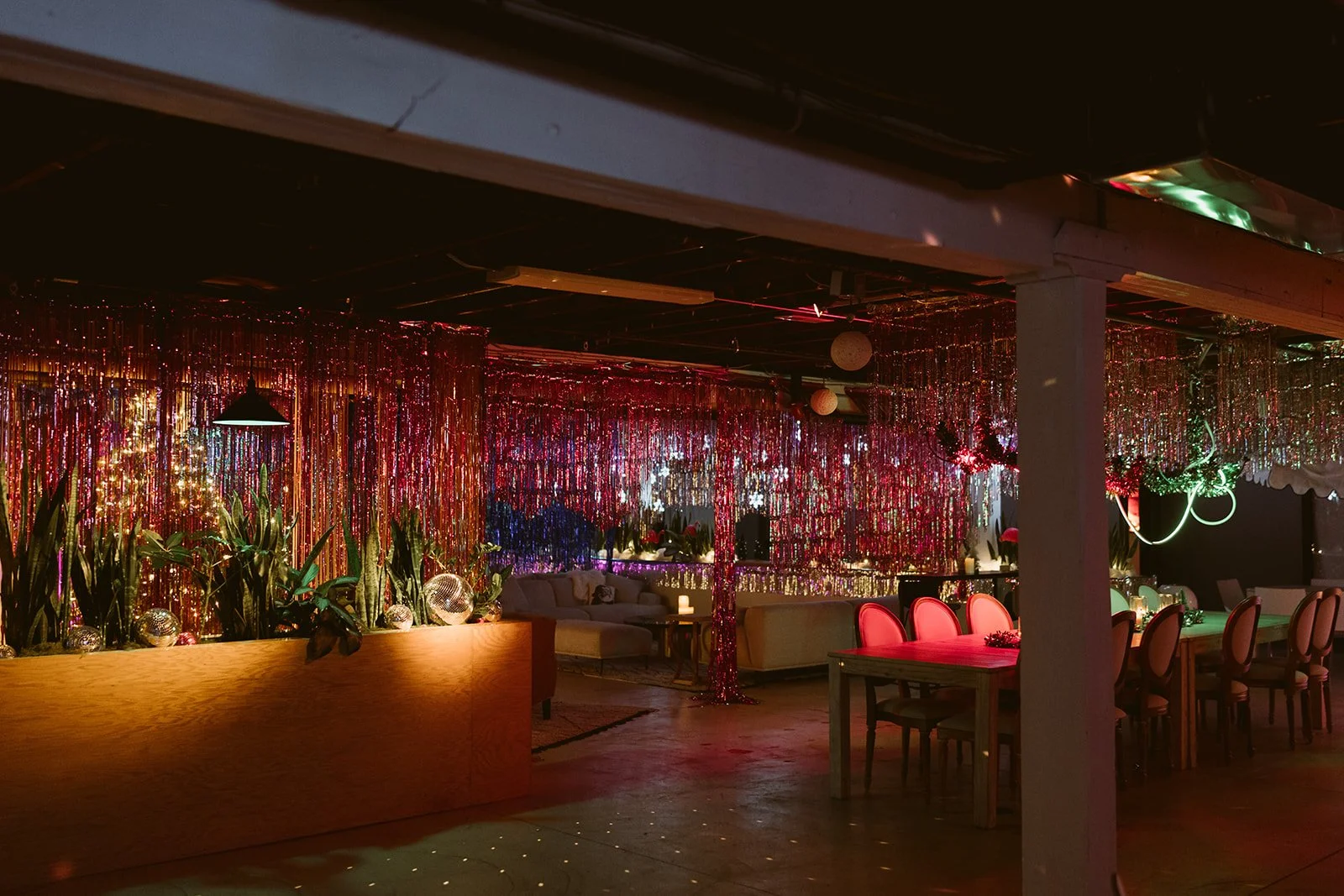 A festive indoor party room decorated with multicolored hanging foil streamers, string lights, and various plants. There are seating areas with couches and chairs, and a table with pink seating.
