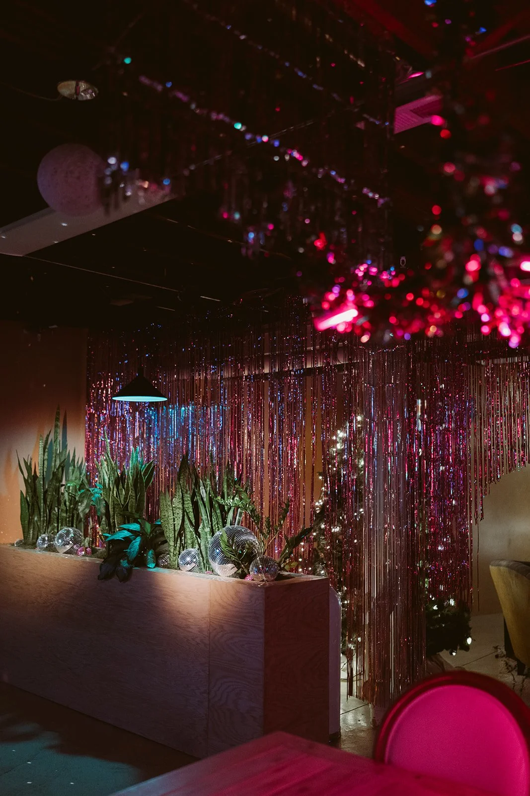 Indoor space decorated with pink and purple glittering streamers, green potted plants, disco balls, and a Christmas tree with lights. Dim lighting enhances the festive atmosphere.