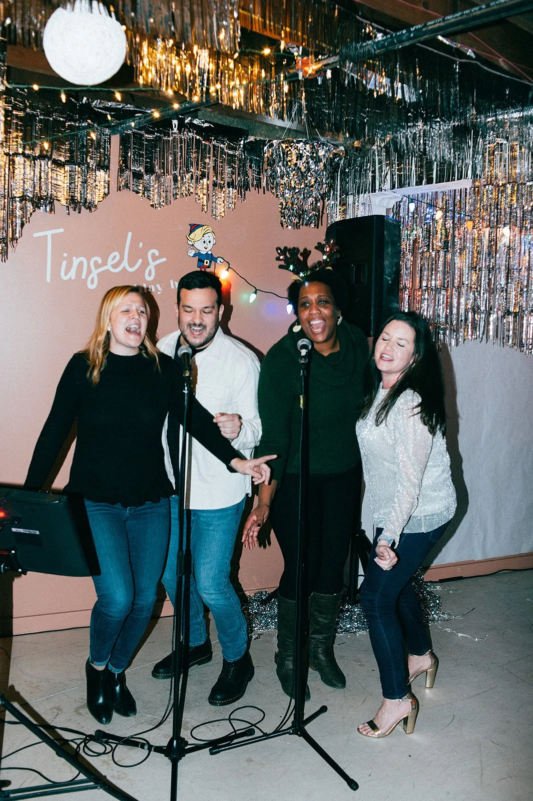 Four people singing or performing together with microphones in a decorated indoor setting with tinsel and a sign that says 'Tinsel's' and a cartoon character of an elf.