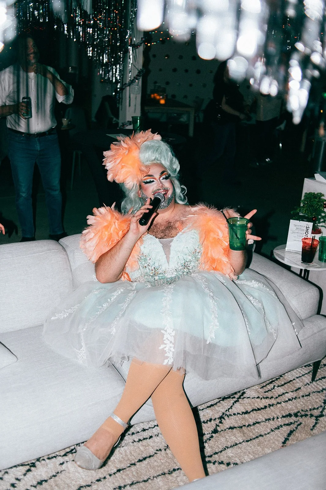 A drag queen in a white dress with orange feathered accents and a large matching hat, sitting on a white couch, holding a microphone and a green drink, smiling at a party or celebration.