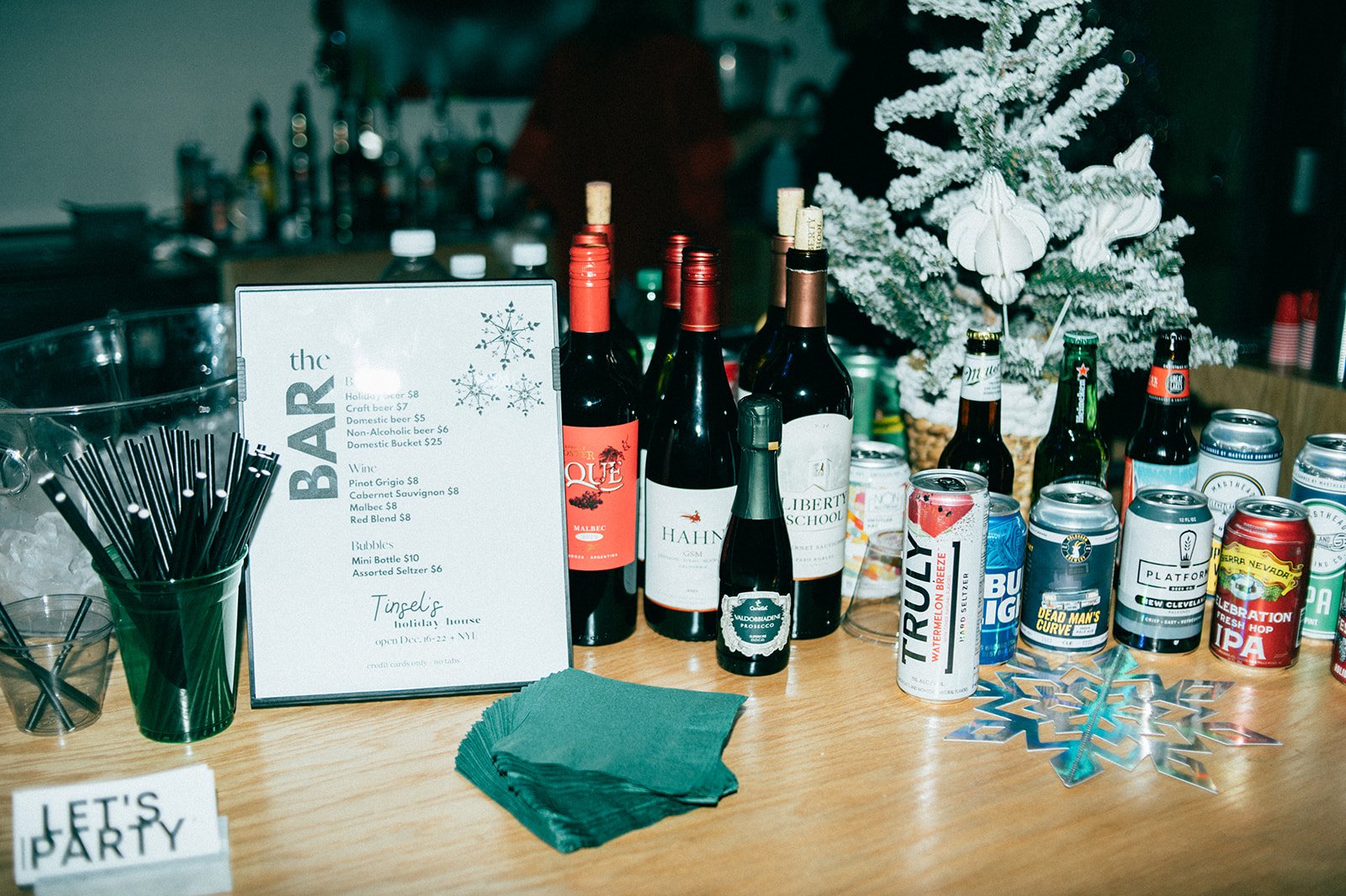 Bar setup with bottled wines, canned beverages, a menu, cocktail straws, napkins, and holiday decorations including a snow-covered tree and snowflake ornaments.