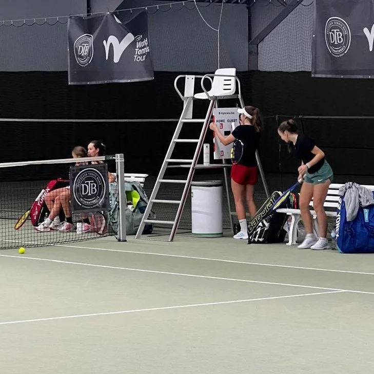 A female tennis player updates the score on MyScoreLive during a tournament in Berlin in Autumn 2025. Two young girls with tennis equipment sit on a bench on an indoor tennis court.