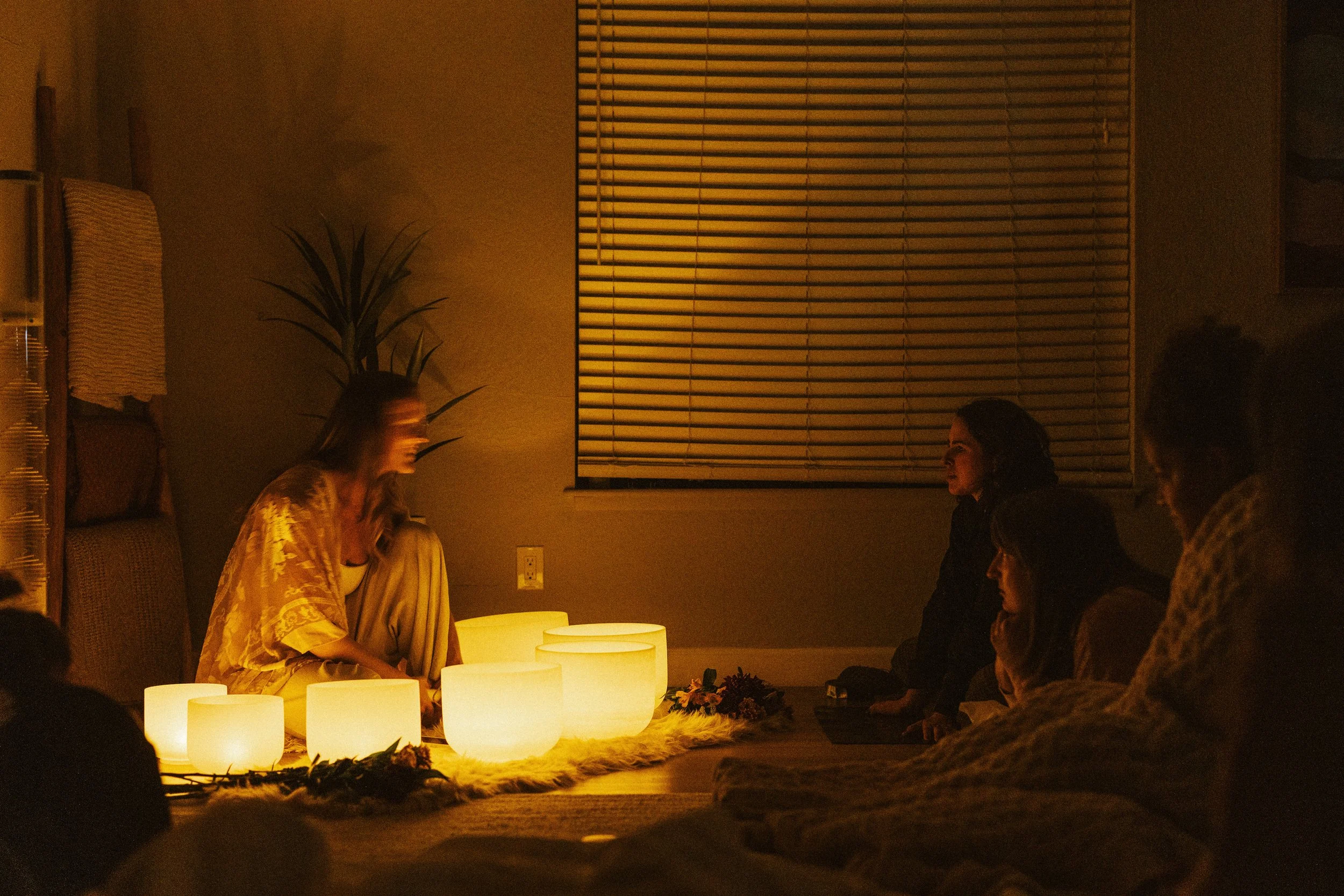 A group of people sitting on the floor in a dimly lit room, facing a woman who is sitting in front of glowing lanterns, with a large window and blinds behind her.