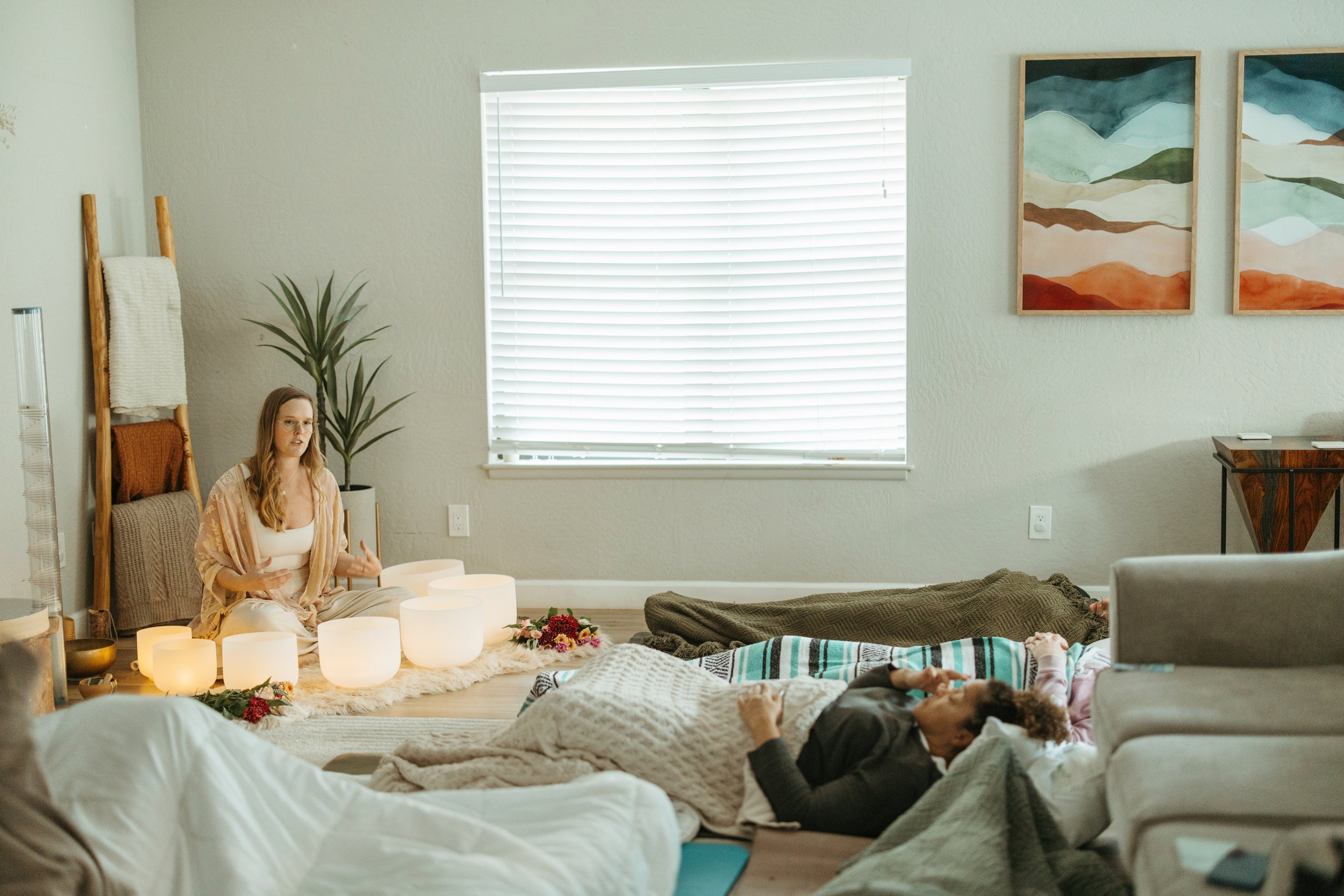 A woman with long hair and glasses sits cross-legged on the floor, speaking, with softly glowing lights around her; two people are lying on the floor, one covered with a blanket, in a cozy living room.