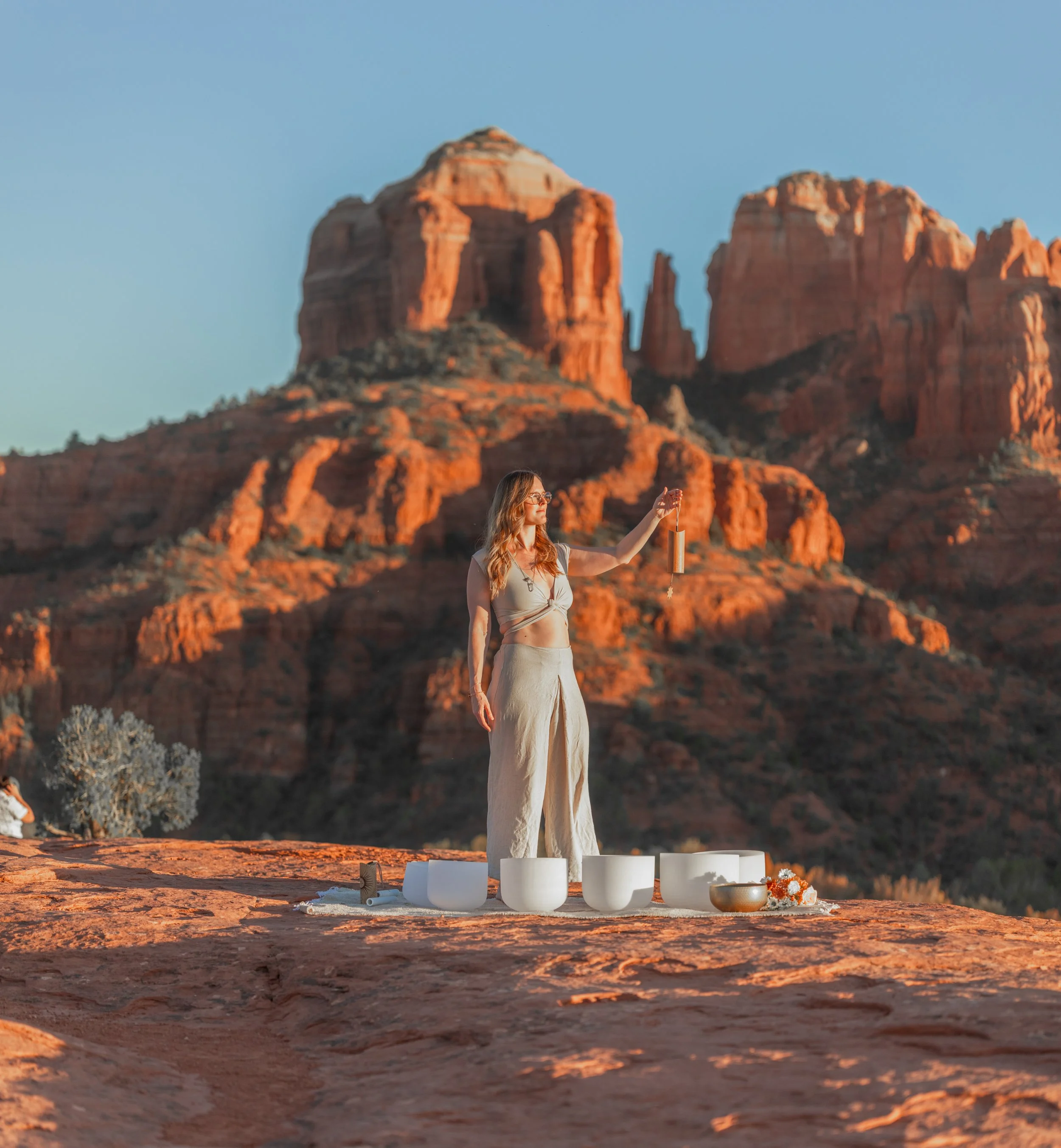 Woman standing outdoors in a desert landscape with red rock formations, holding a wind chime, surrounded by singing bowls and flowers.