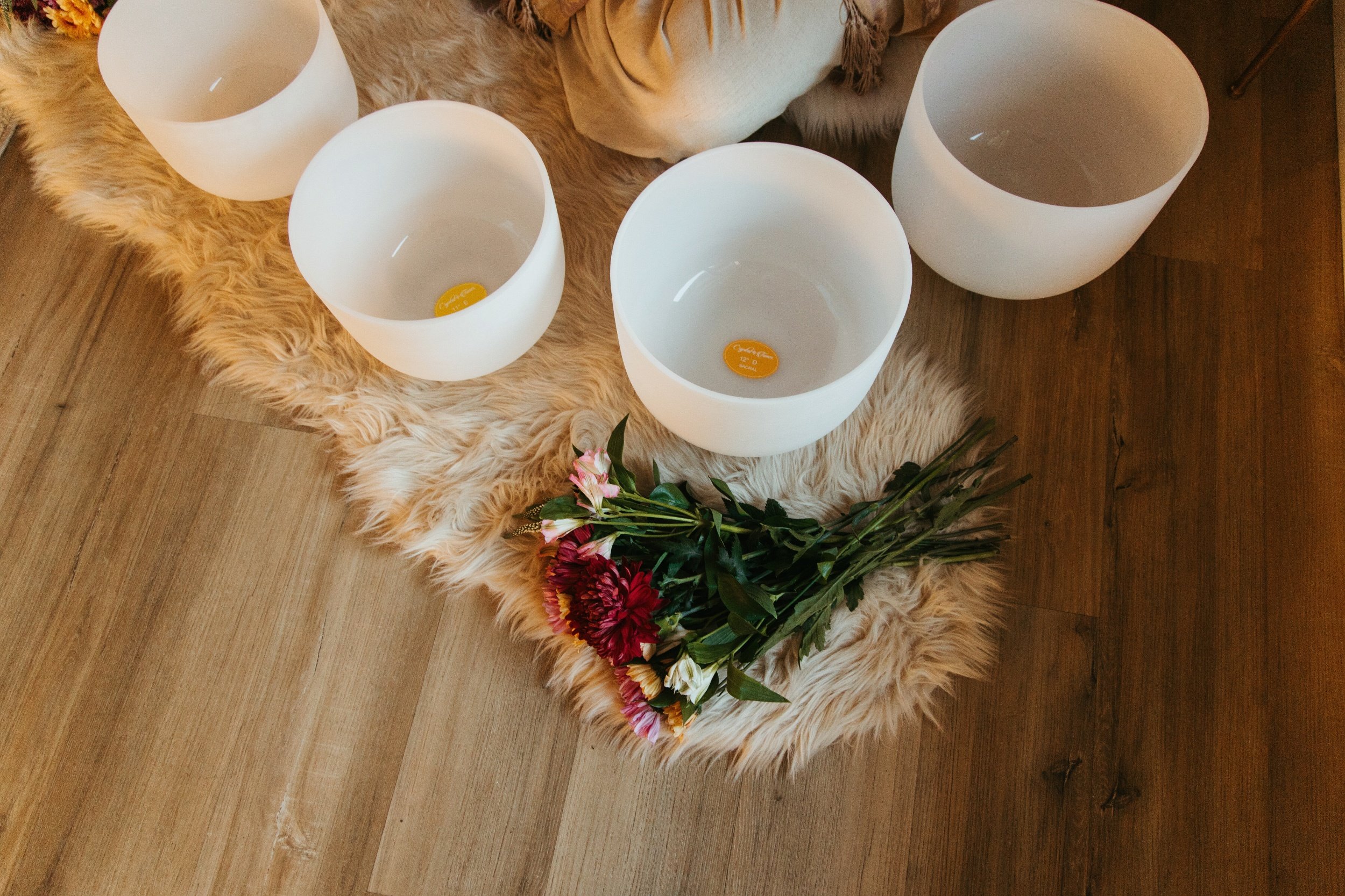Four white singing bowls on a cream-colored furry rug with a bouquet of pink, red, and white flowers placed on the rug, on a wooden floor.