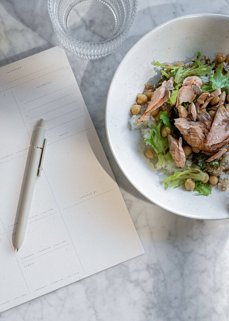 A marble table with a glass of water, a blank planner with a white pen, and a white bowl containing salad with chickpeas, greens, rice, and shredded meat.