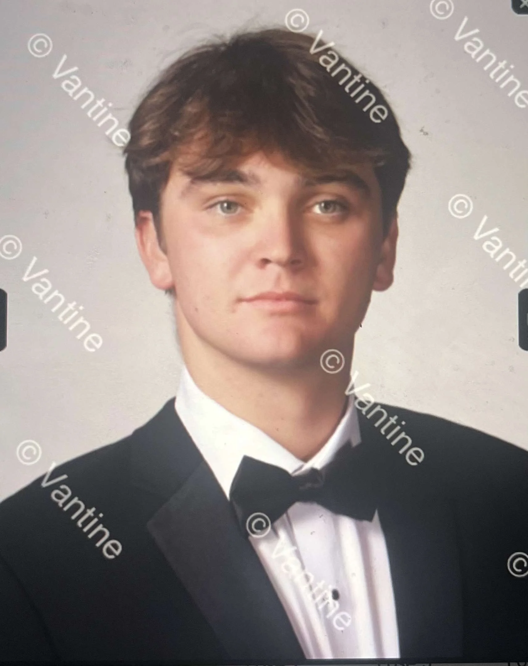 Young man in formal attire wearing a black tuxedo jacket, white shirt, and black bow tie, smiling in a portrait-style photo.