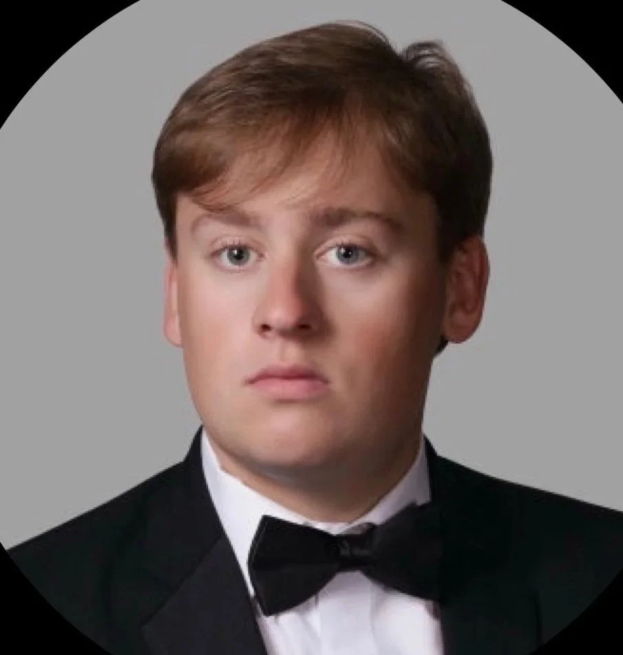 Young man wearing formal attire, smiling, with a crest in the background.