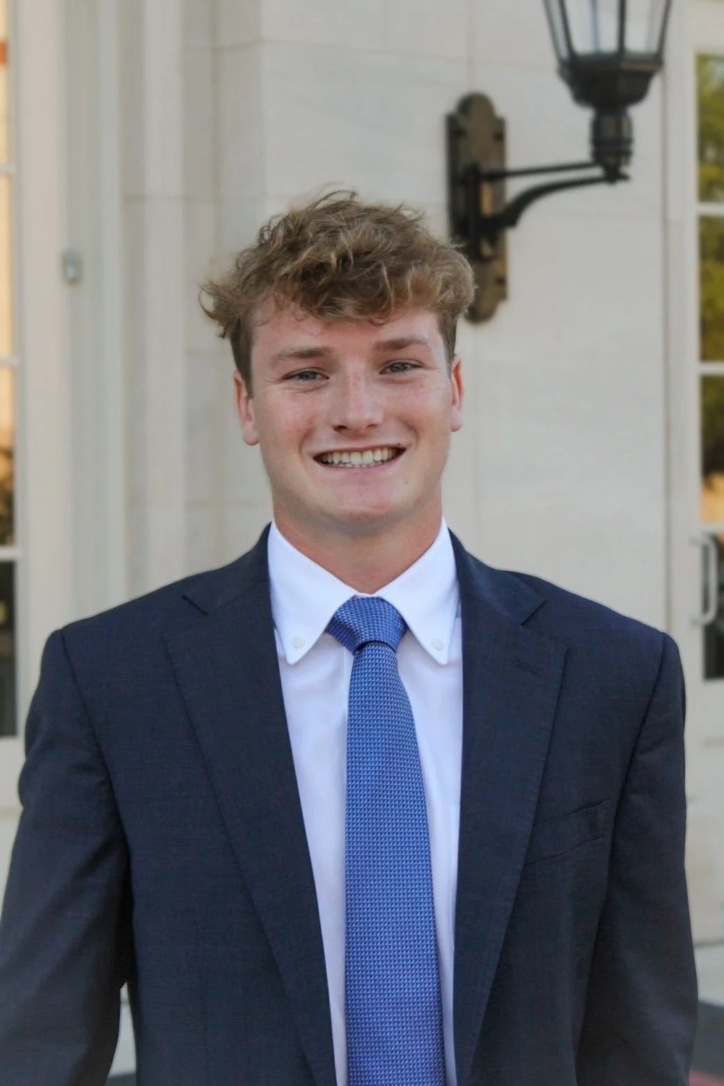 A young man with curly hair smiling, wearing a suit jacket and shirt. Indoors with a framed photo in the background.