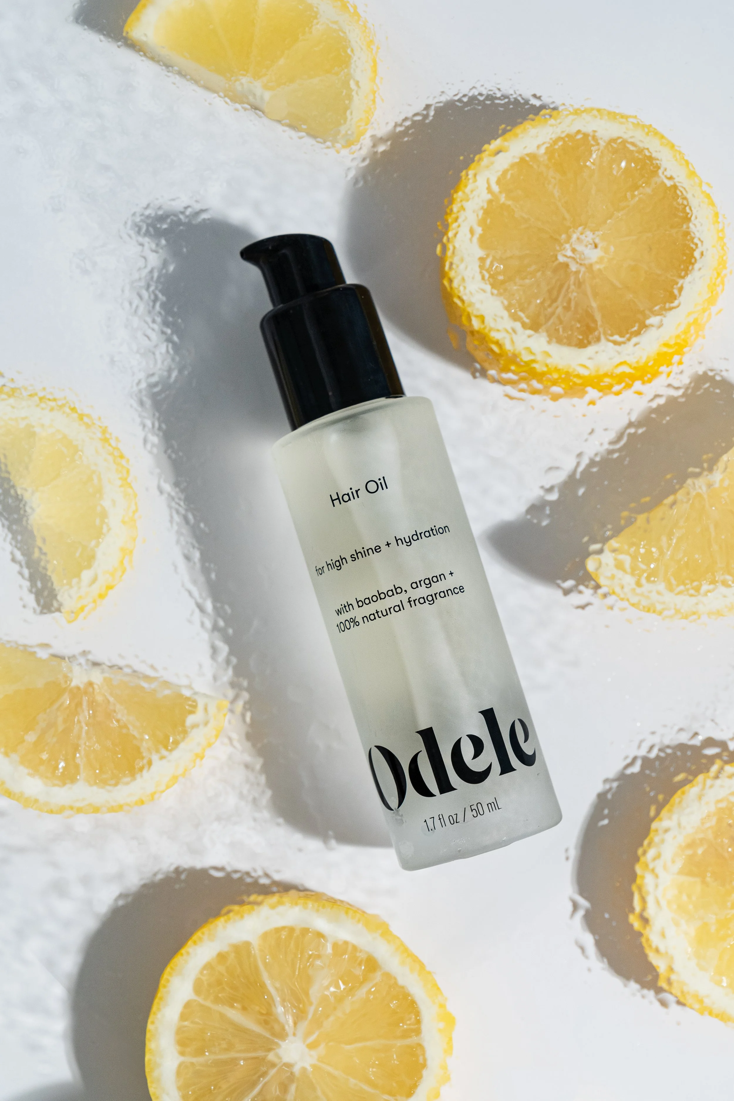 A frosted white bottle of hair oil with a black pump dispenser labeled 'Odele' is surrounded by sliced lemons on a textured surface.