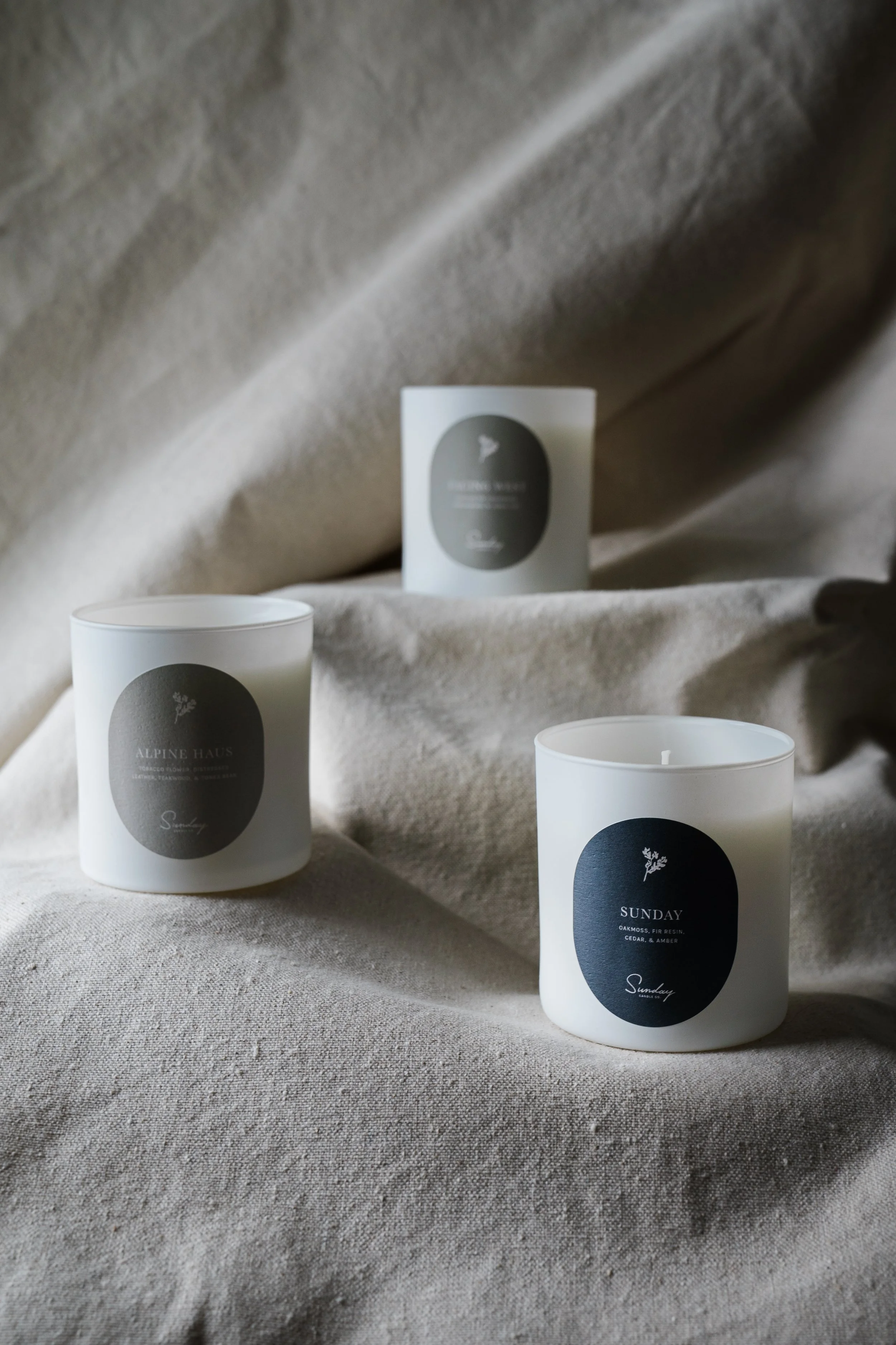 Three white candles with black and gray labels on a beige fabric background.