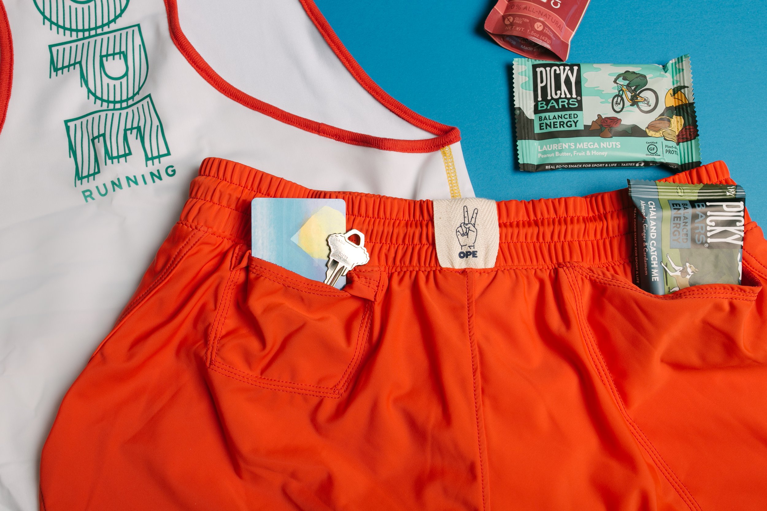 Flat lay of red running shorts, a white and red tank top, and energy snacks on a blue background. The tank top has the words 'U RUNNING' with a stylized mountain graphic. The shorts have a key and an OPE symbol attached, with the pocket containing a 