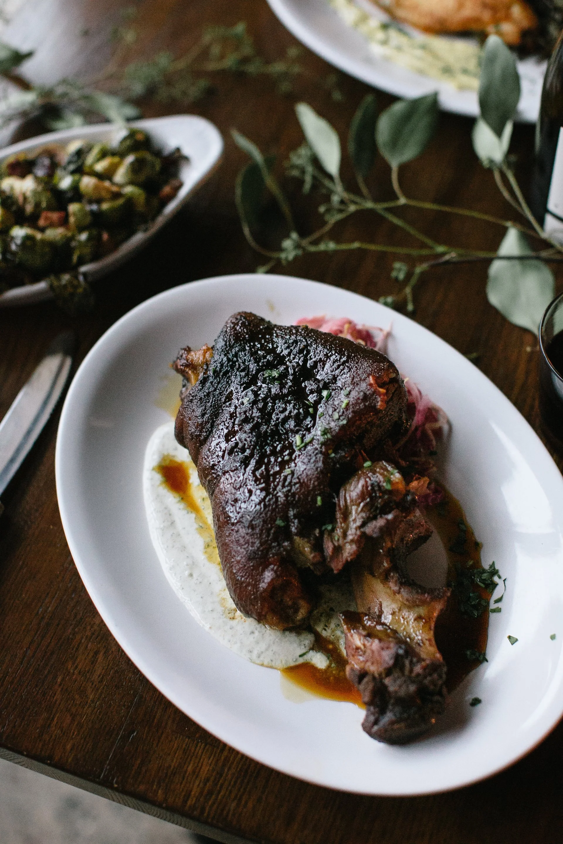 A plated cooked beef short rib with dark sauce on top, served on a white oval plate with sides of sauce and garnish. Additional dishes and a glass of red wine are visible on the table.