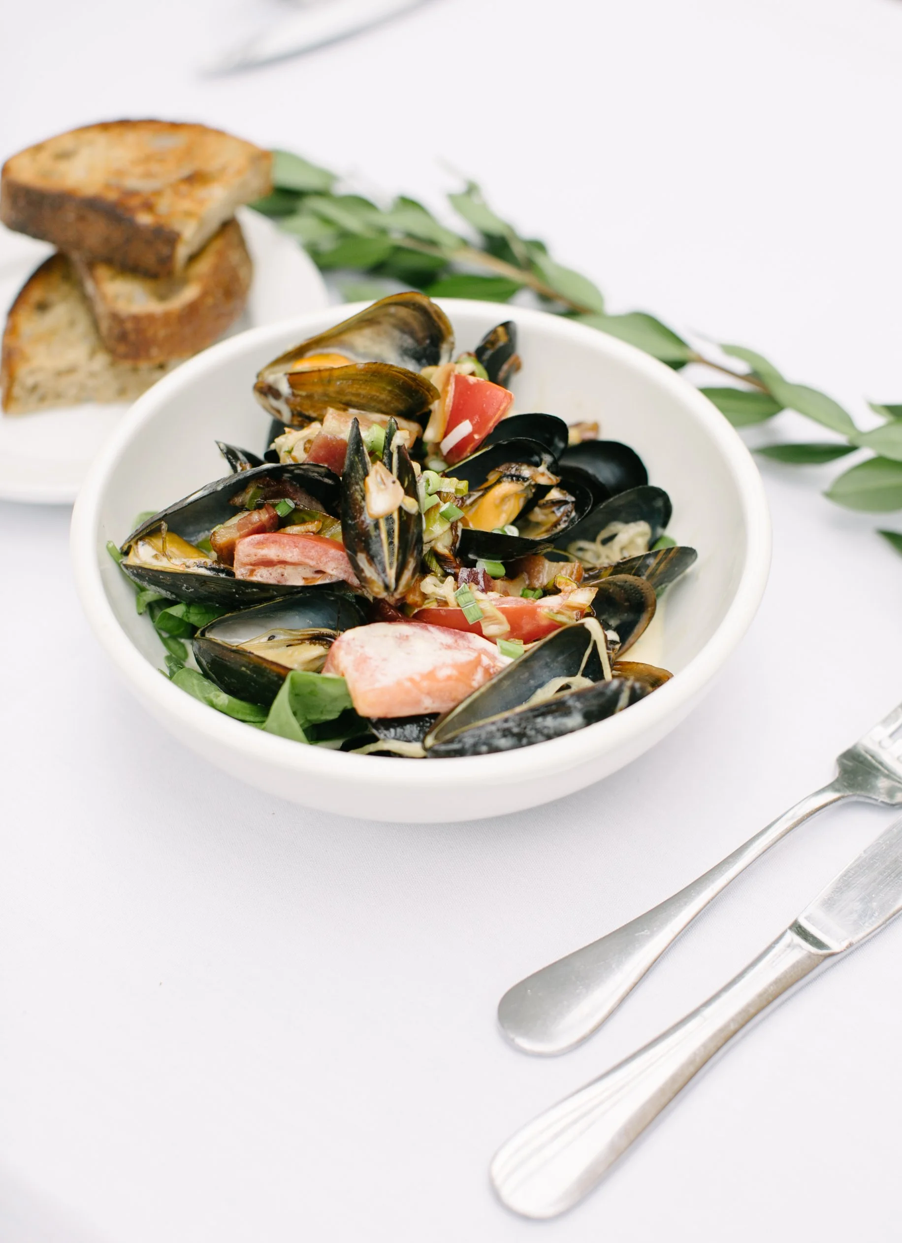 Bowl of cooked mussels with chopped vegetables and greens, with slices of toasted bread on a small plate, on a white table with a fork and knife alongside.