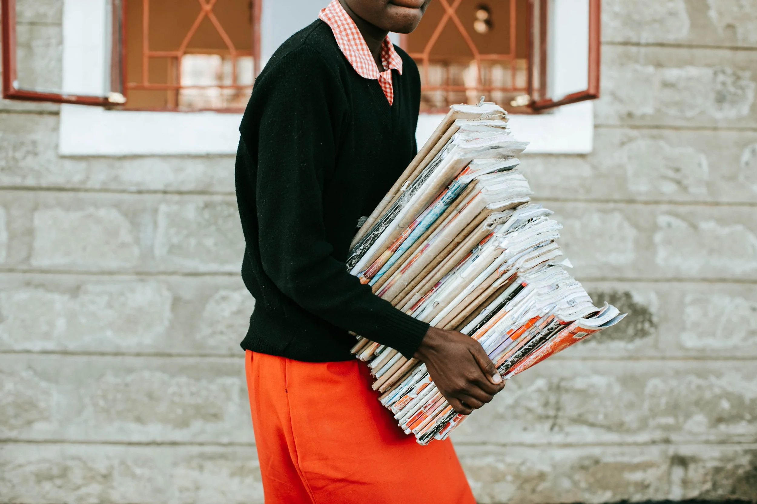 Person holding a large stack of newspapers outdoors in front of a stone wall with open windows.