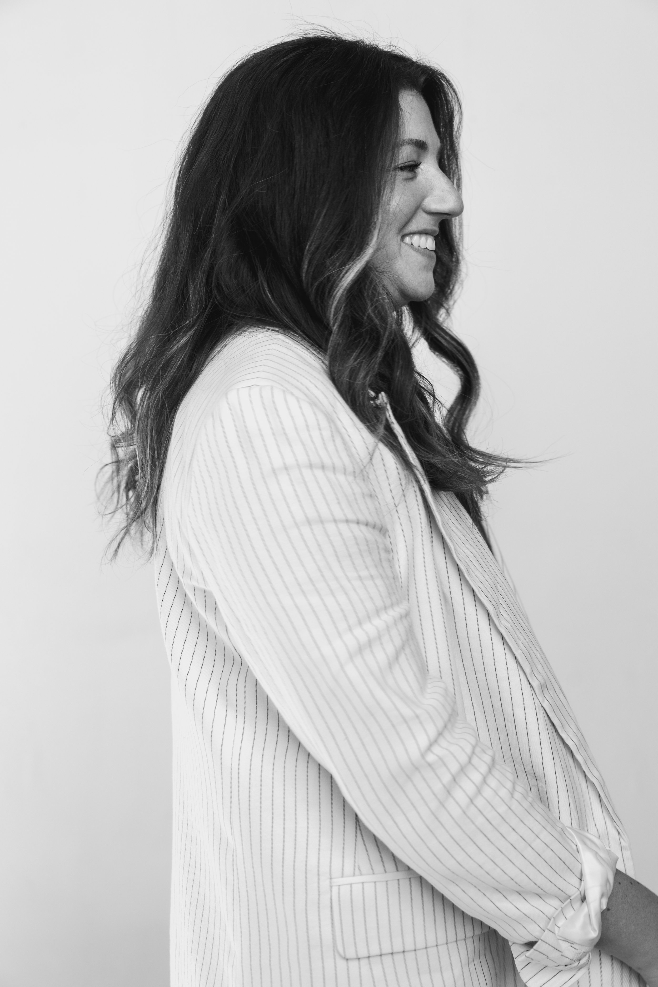 Black and white photo of a woman with long wavy hair, smiling, wearing a striped blazer, facing right.