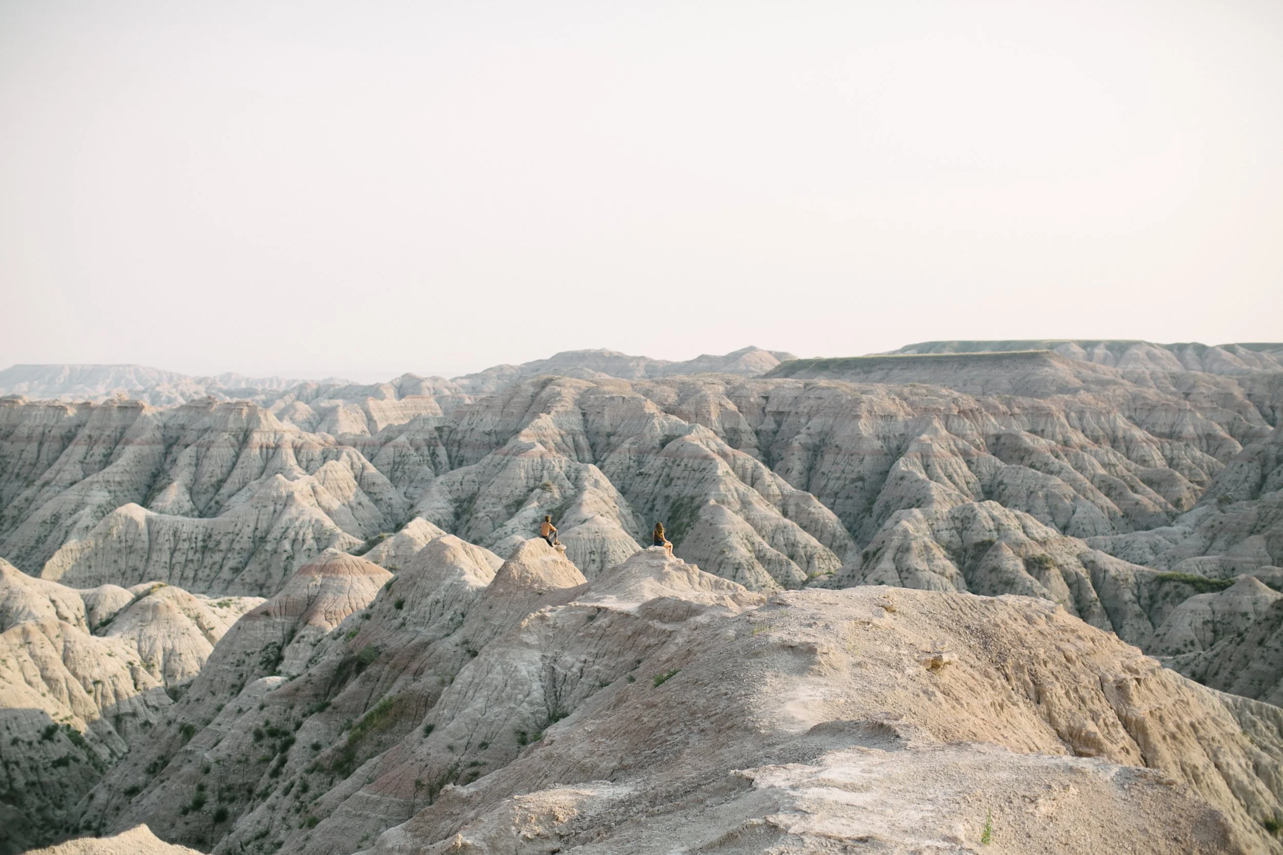 Two people sitting on rocks overlooking rugged, eroded badlands landscape with layered rock formations under a clear sky.
