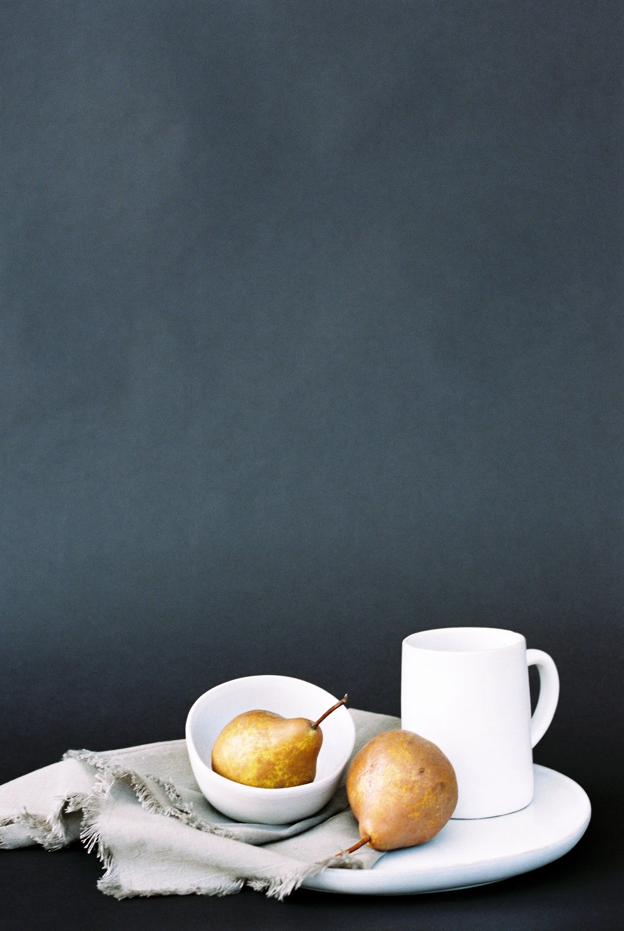 Two pears in a white bowl, one empty white mug, and a small round plate on a beige cloth against a dark background.