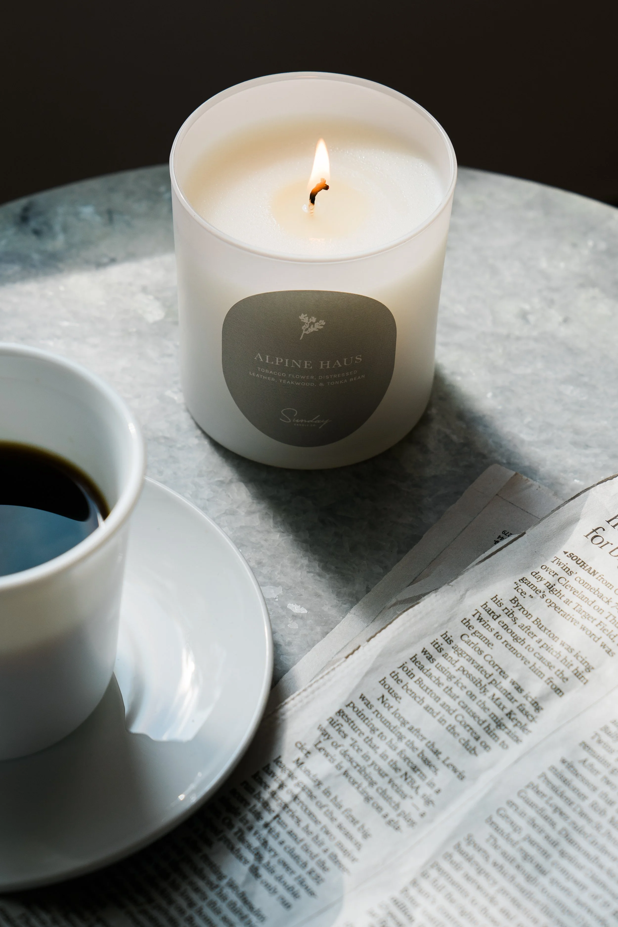 A white candle labeled 'Alpine Haus' burning on a marble surface, a cup of black coffee on a saucer, and an open newspaper.