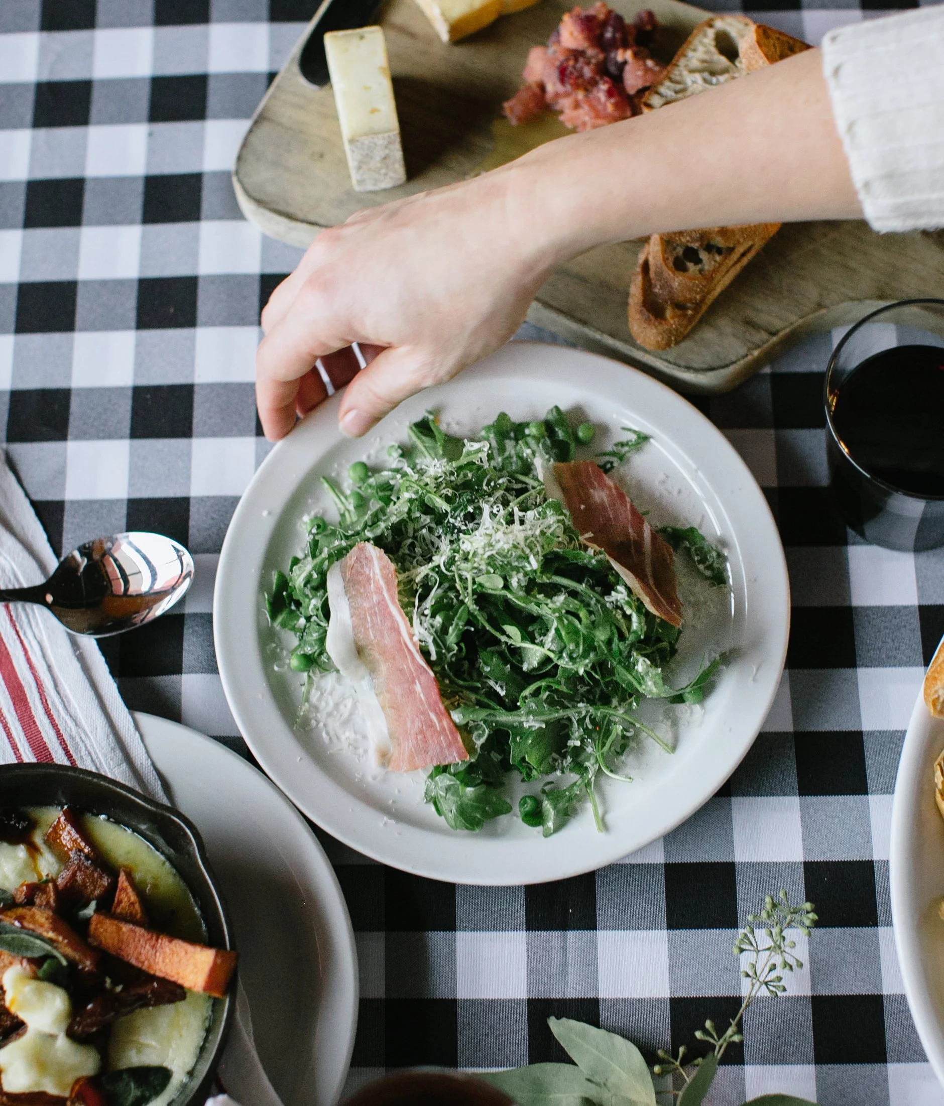 A hand reaching over a plate of arugula salad with prosciutto, grated cheese, and a glass of red wine on a checkered tablecloth.