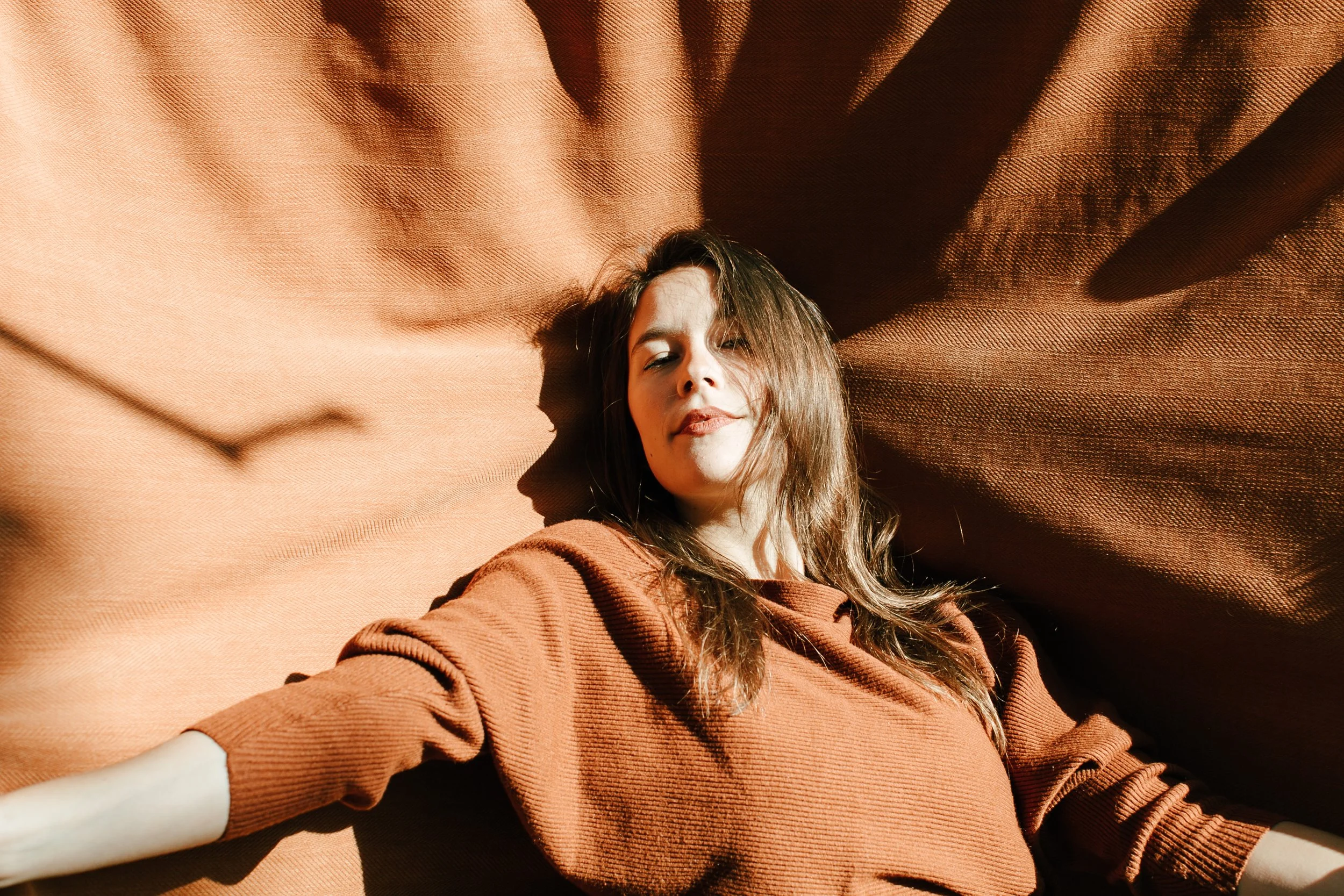 A woman with brown hair lying on a textured red or orange fabric surface, with sunlight casting shadows on her face and the fabric.