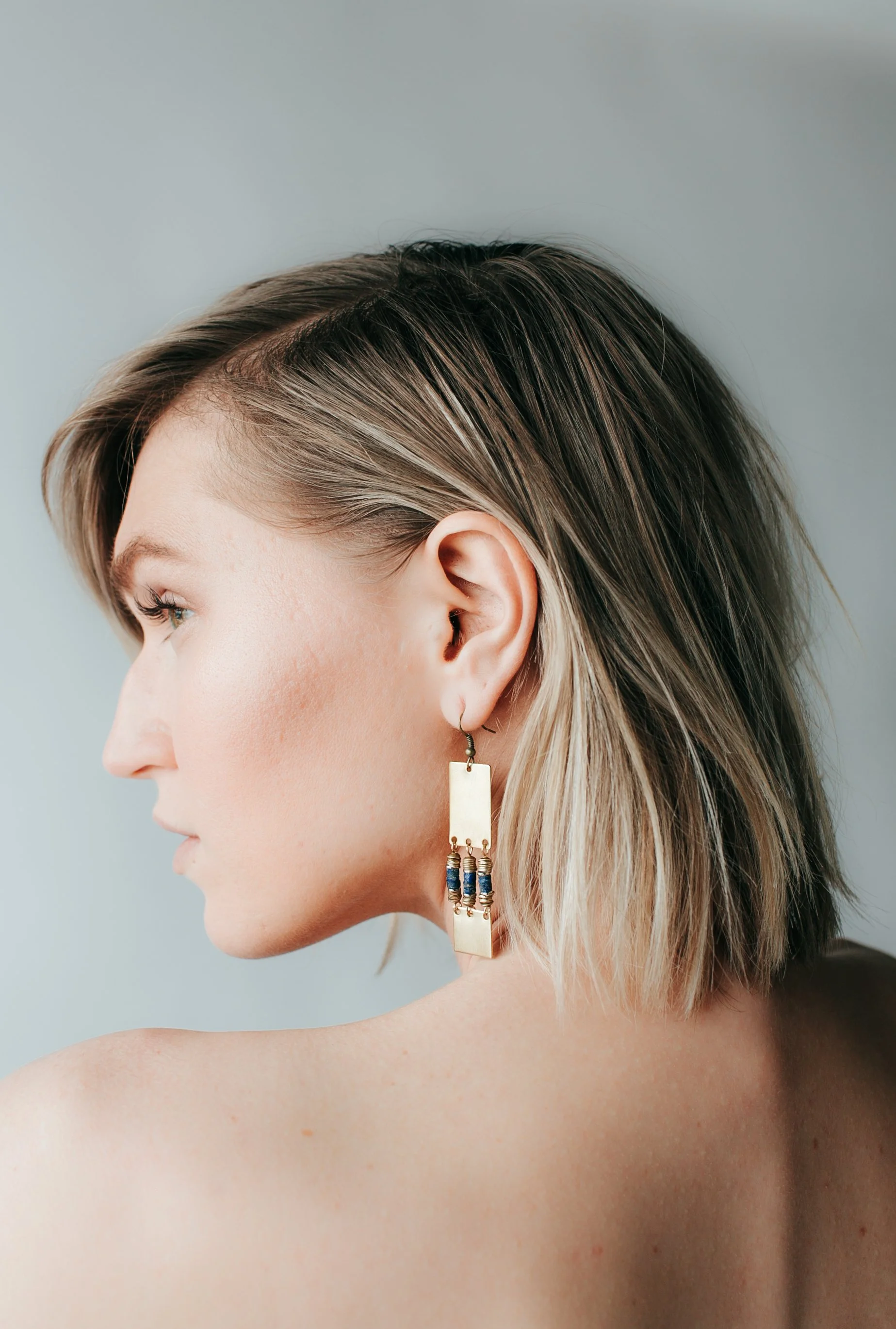 Profile of a woman with short, layered, blonde hair, wearing a dangling gold earring with beads, against a light-colored background.