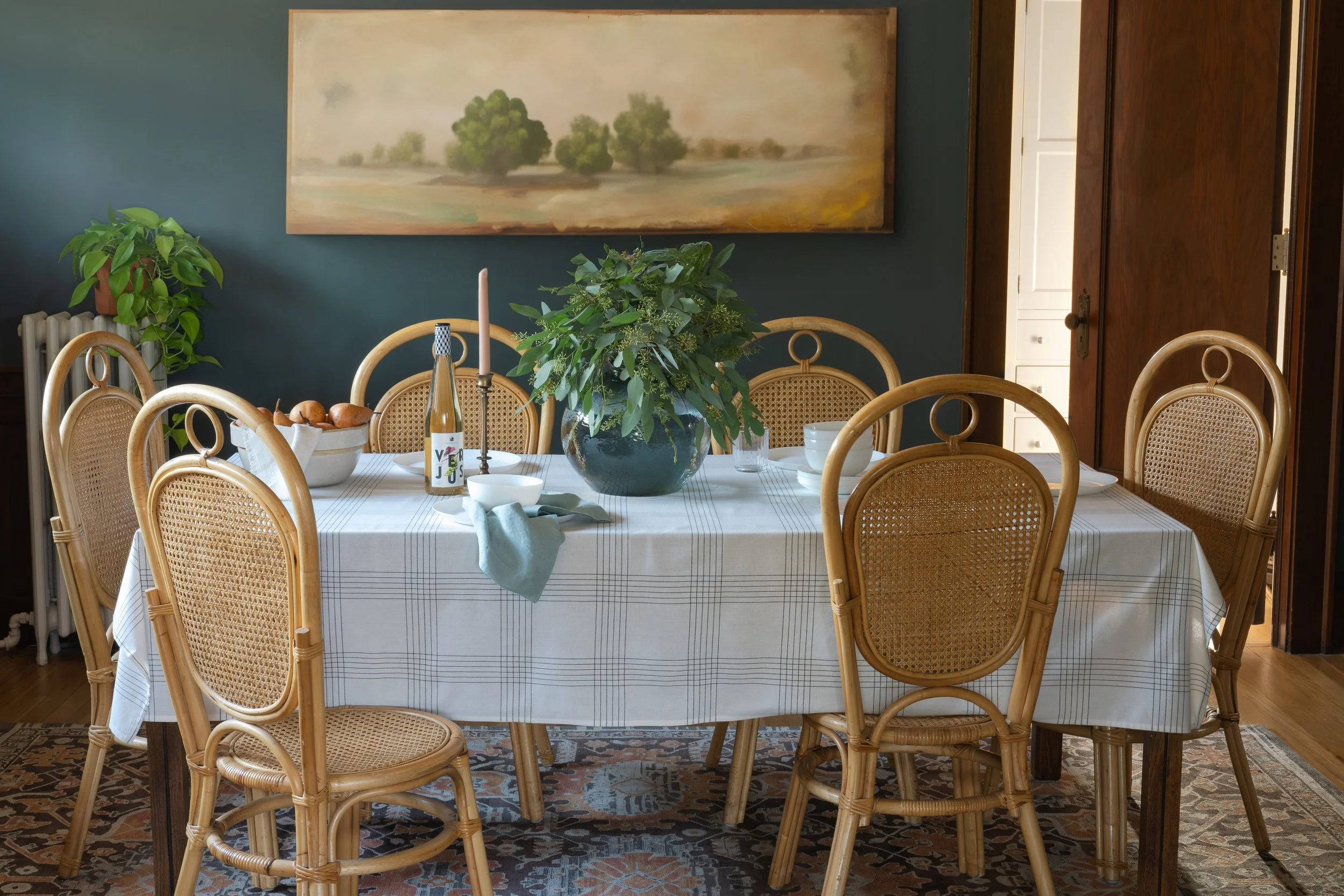Dining room with a table set with bowls, a bottle, a candle, and a vase of greenery, surrounded by bamboo-style chairs, with a large landscape painting on the dark green wall.