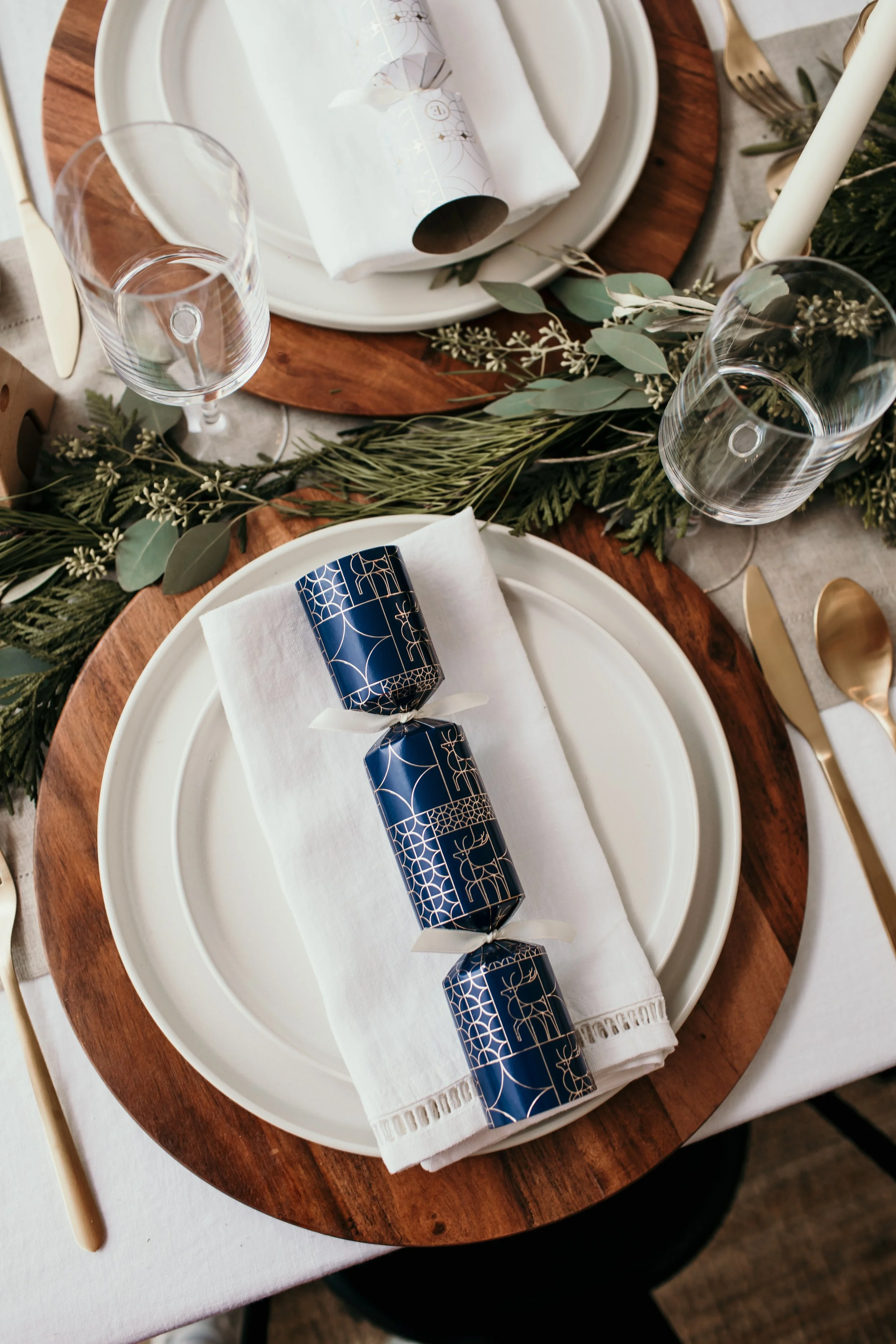Elegant holiday table setting with white plate, white cloth napkin, decorative blue and gold wrapped candies, greenery garland, wine glass, and gold utensils.