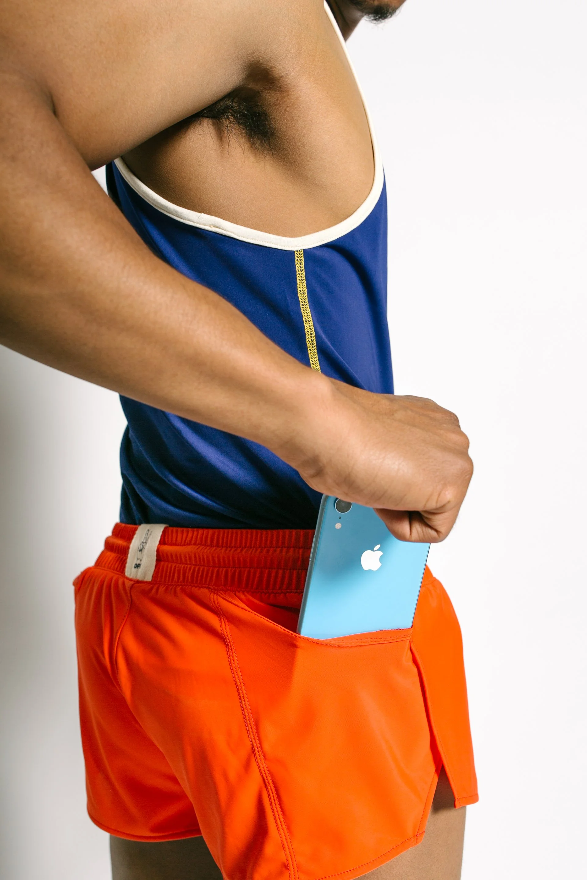 A person wearing an orange running shorts and a blue athletic shirt with white trim, putting a blue iPhone into their front pocket.