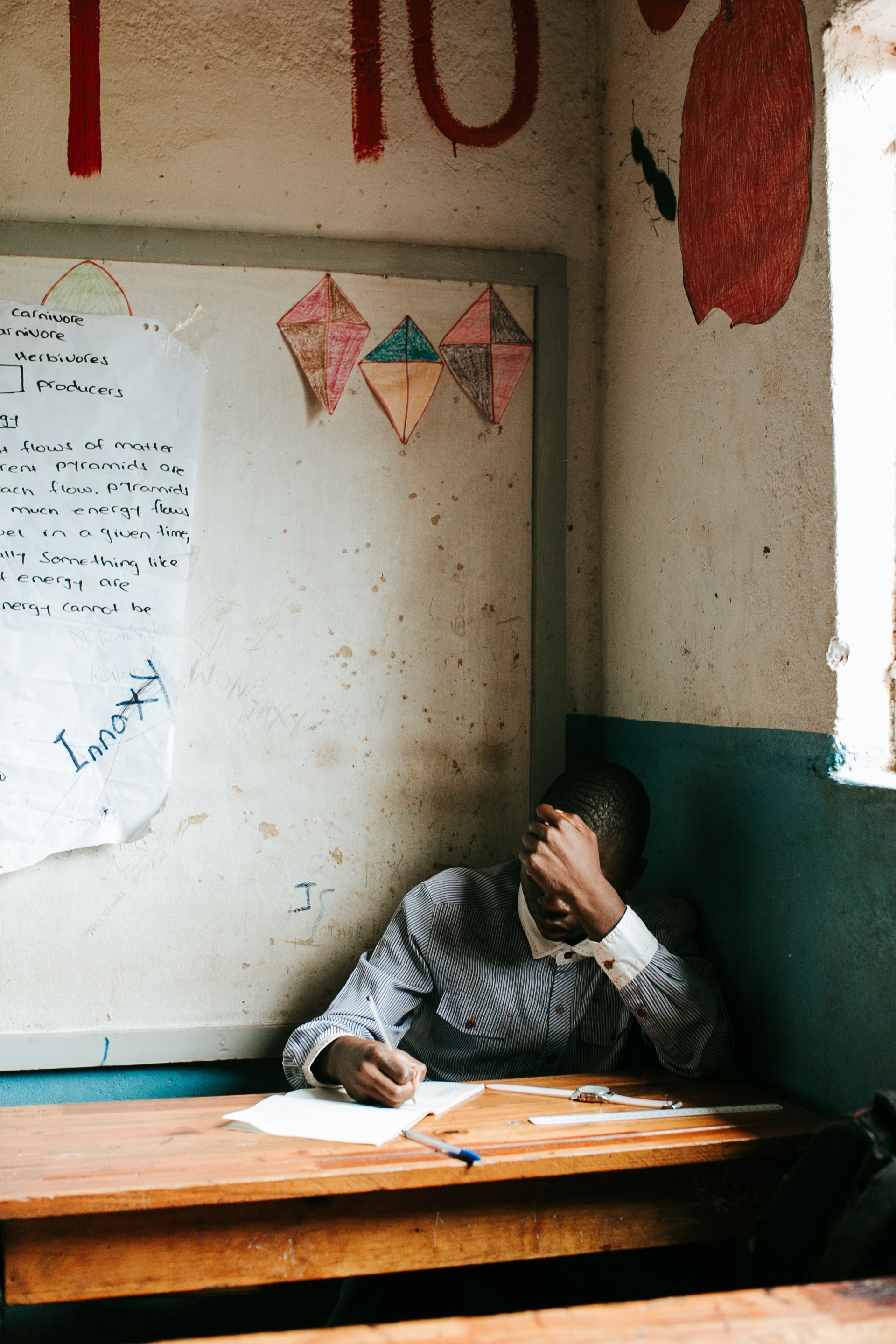 A young man sitting at a desk with his head resting on his hand, in a classroom with a poster and drawn paper lanterns on the wall behind him.