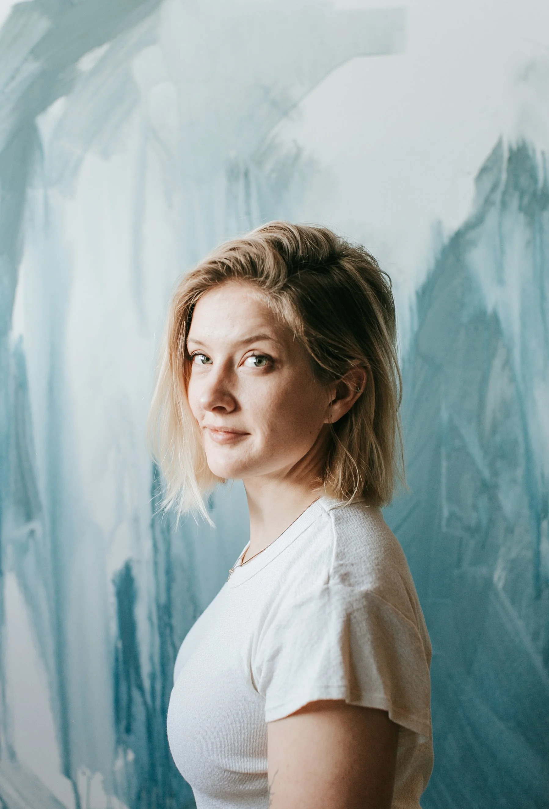 A woman with shoulder-length blond hair and blue eyes standing in front of an abstract blue and white painted background.