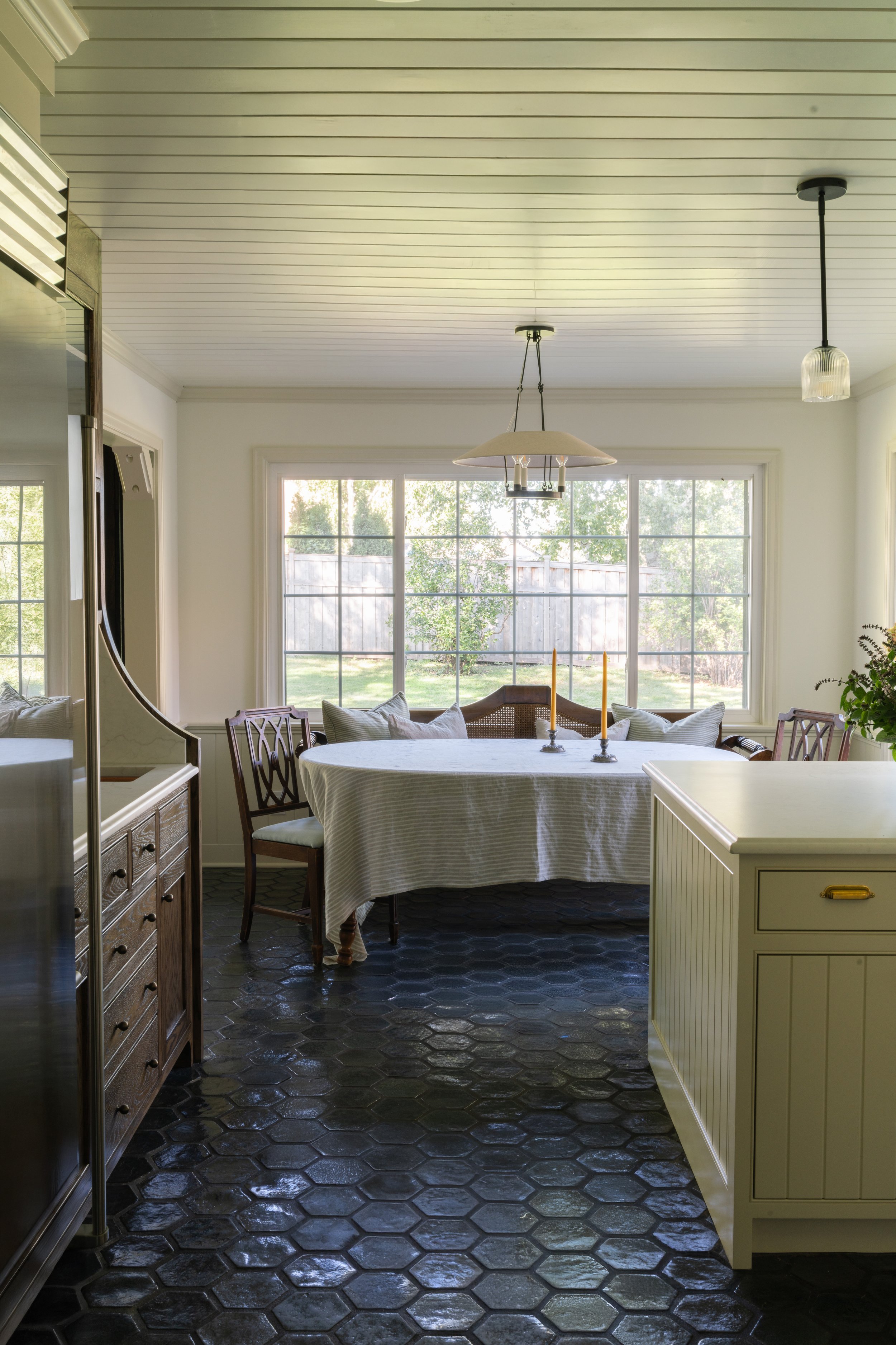 Bright dining room with large window, round table with a white tablecloth, wooden chairs with cushions, and a black hexagonal tiled floor.