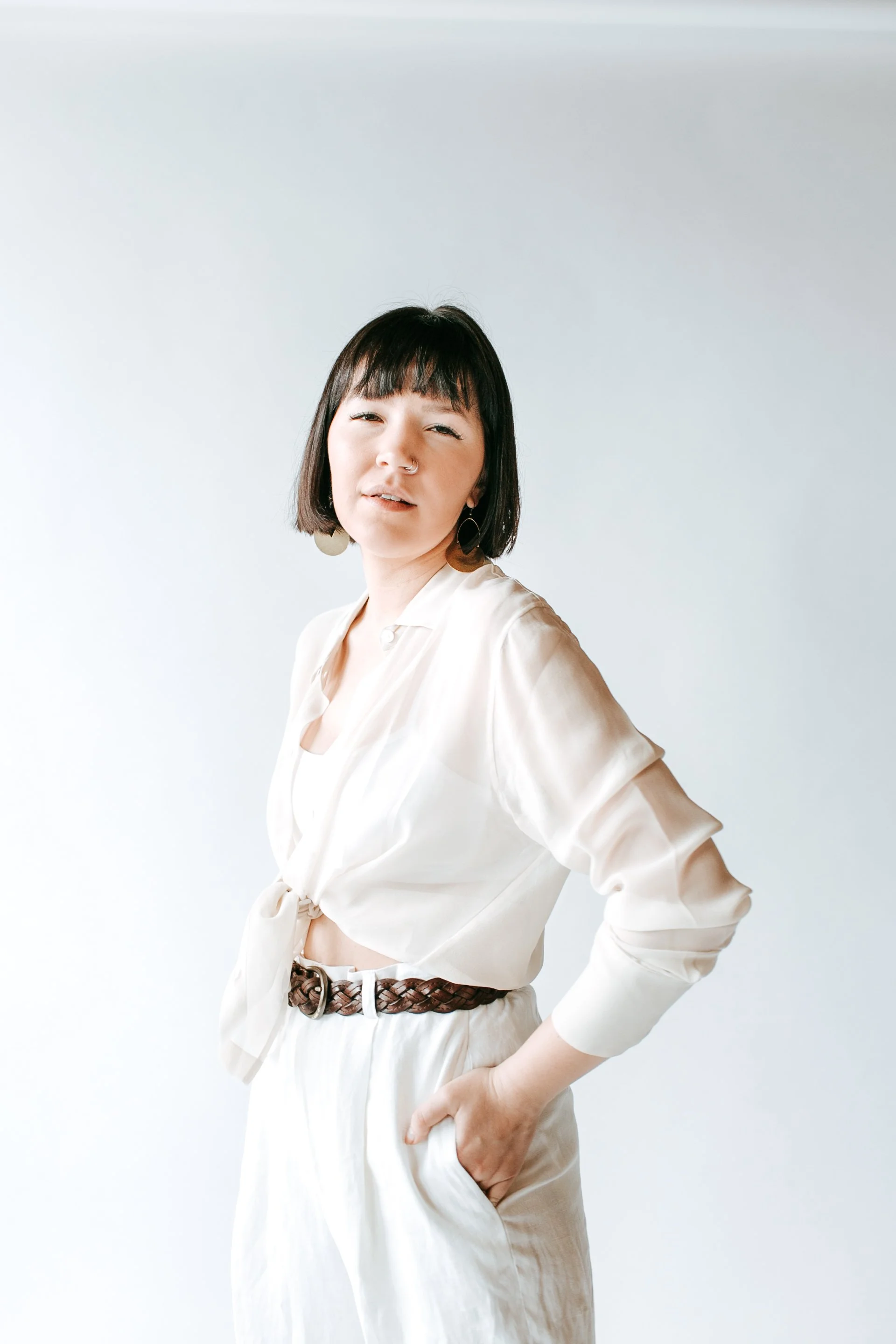Young woman with short dark hair winks and looks at the camera, wearing a cream-colored blouse and white pants with a brown braided belt, standing against a plain white background.