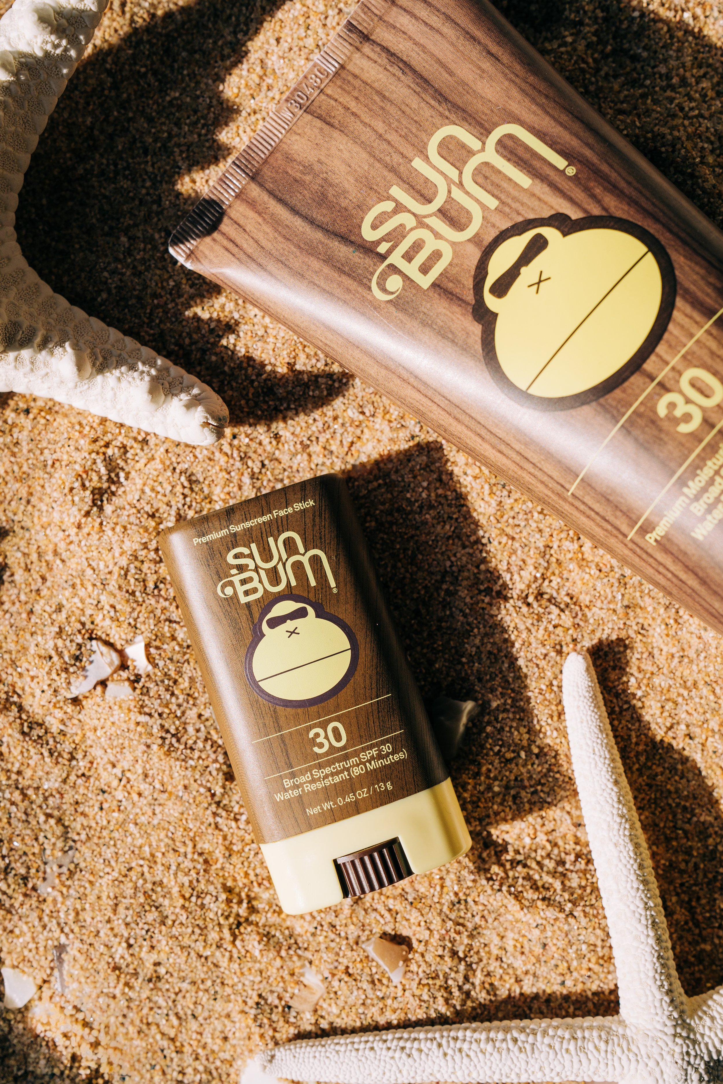 A tube and box of Sun Bum SPF 30 sunscreen lying on sandy beach with a starfish and shell nearby.