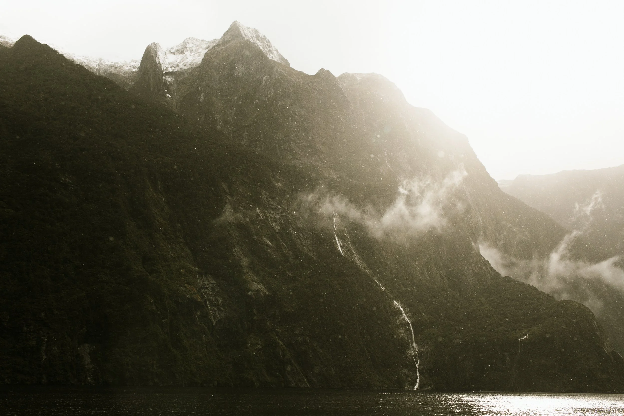 Mountains with waterfalls and clouds over a body of water, sunlight illuminating the scene.