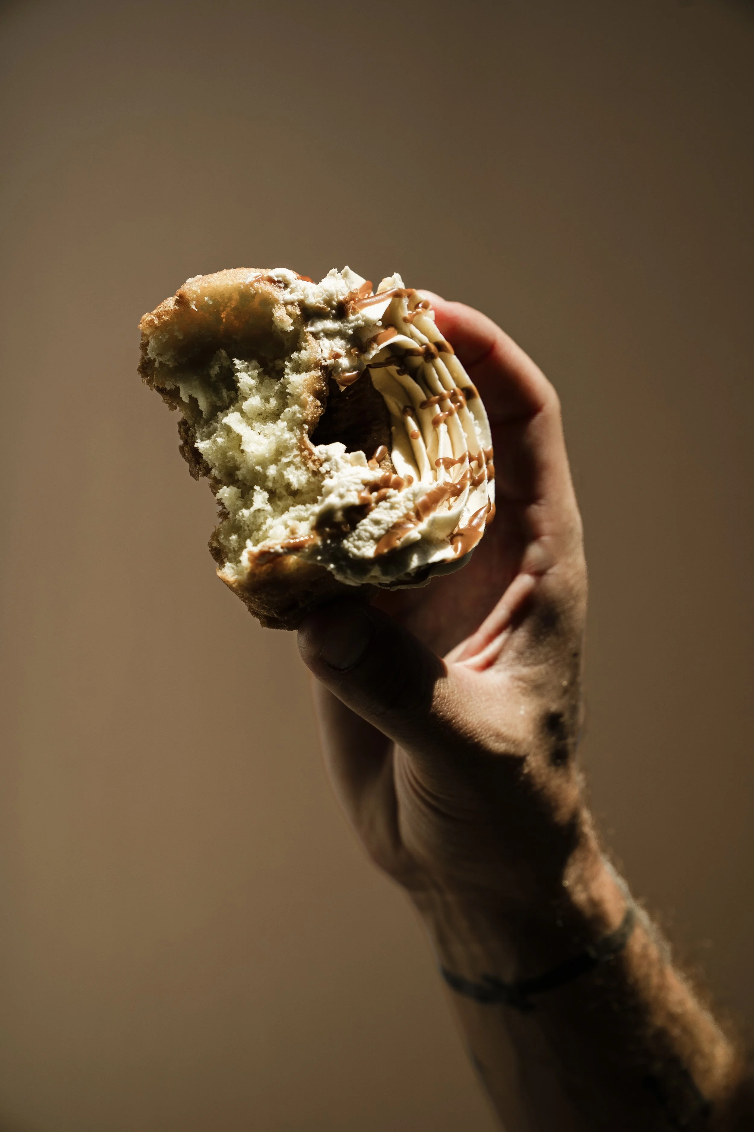 A hand holding a half-eaten chocolate and vanilla ice cream donut with drizzled caramel and chocolate on top, against a plain background.