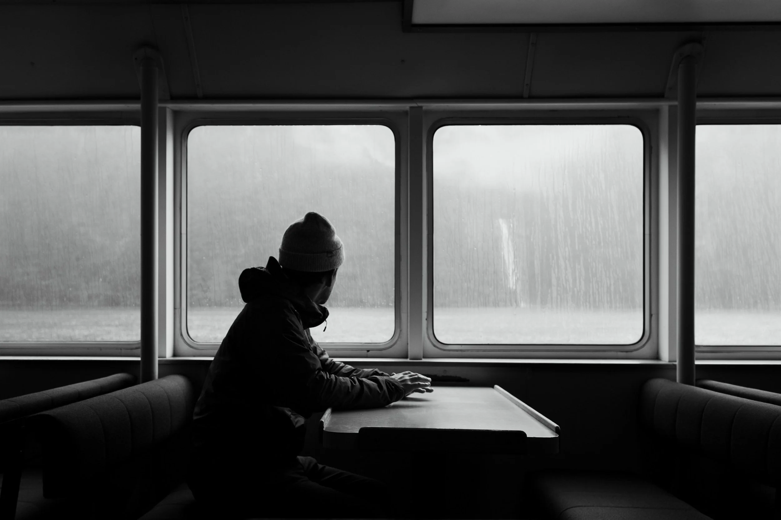 A person sitting alone on a bench inside a ferry or train, gazing out the window on a rainy day.