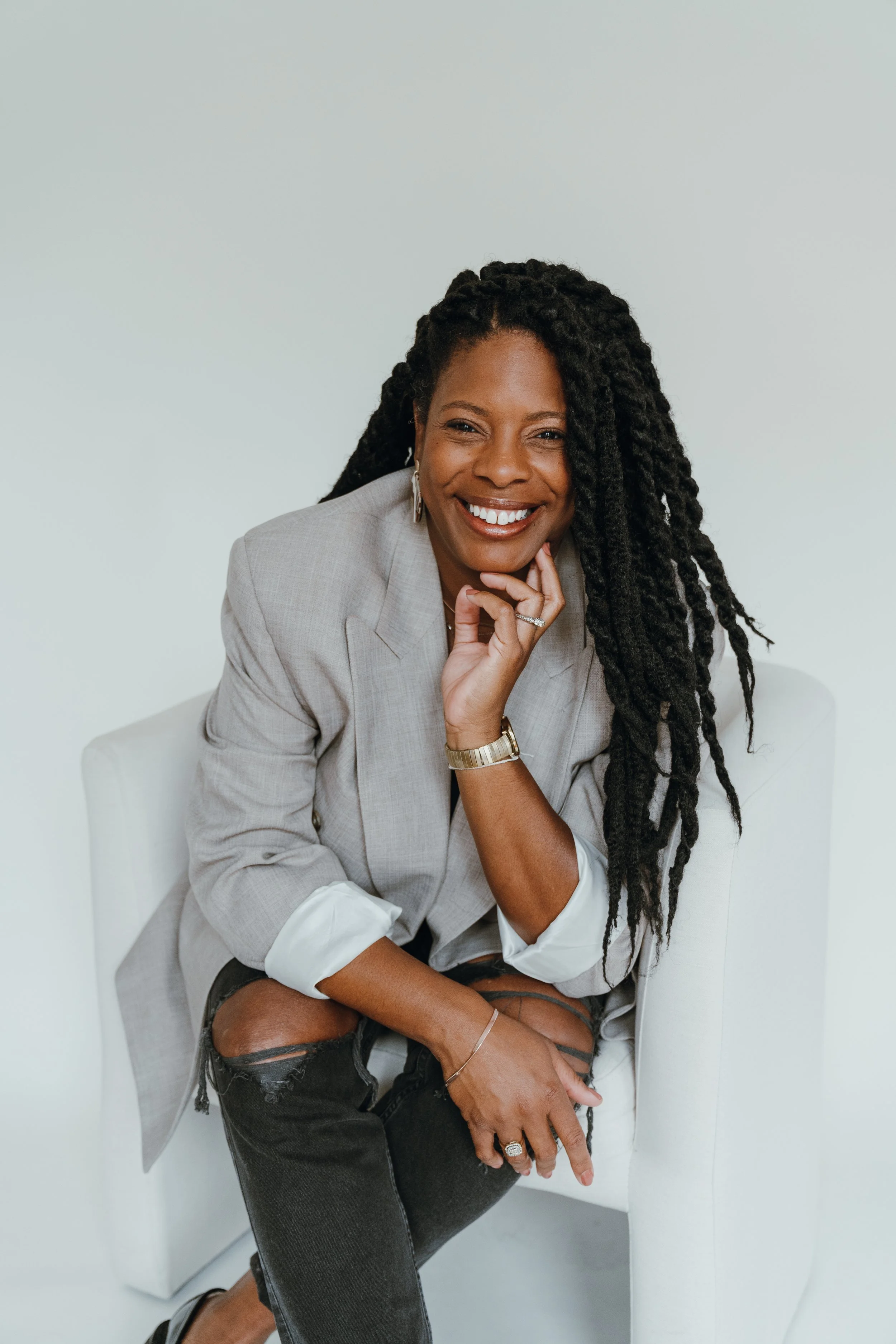 A woman with long dreadlocks smiling, sitting on a white chair, wearing a light gray blazer and distressed black jeans.