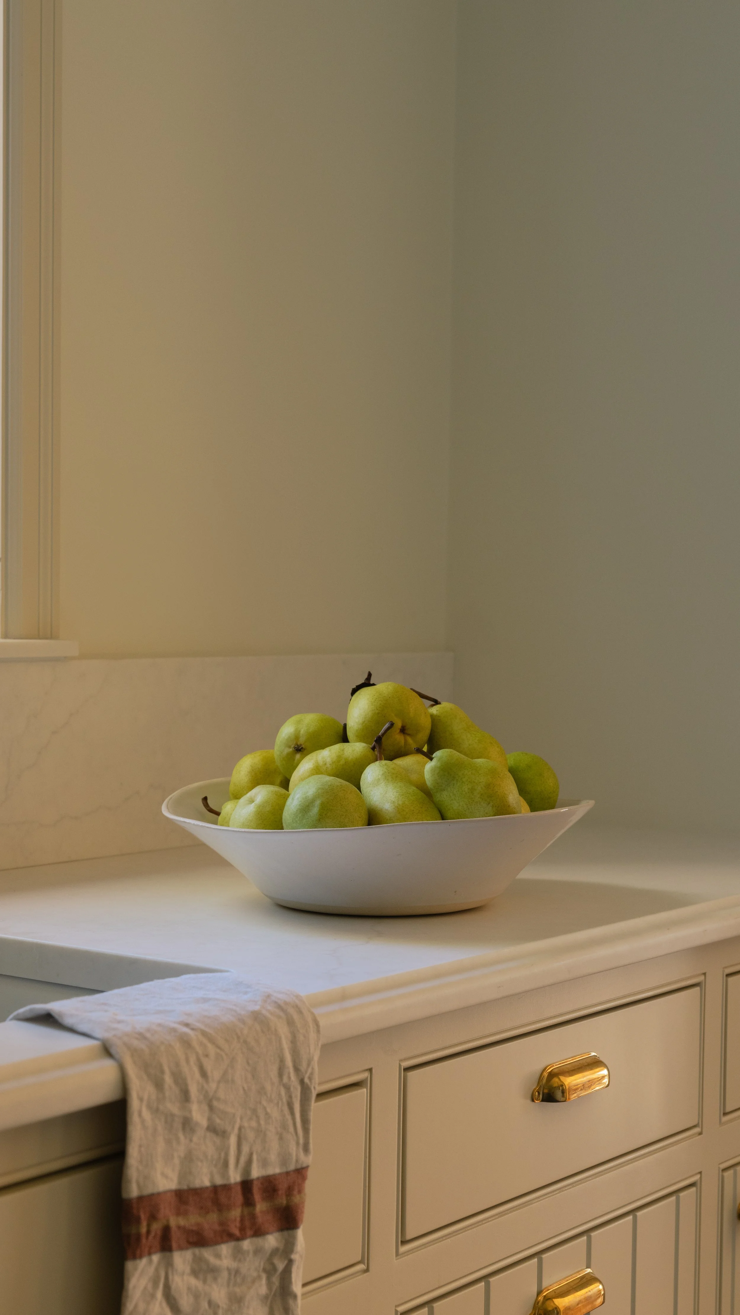 A white bowl filled with green pears on a white marble countertop, with a drawer below and a cloth hanging over the edge of the counter.