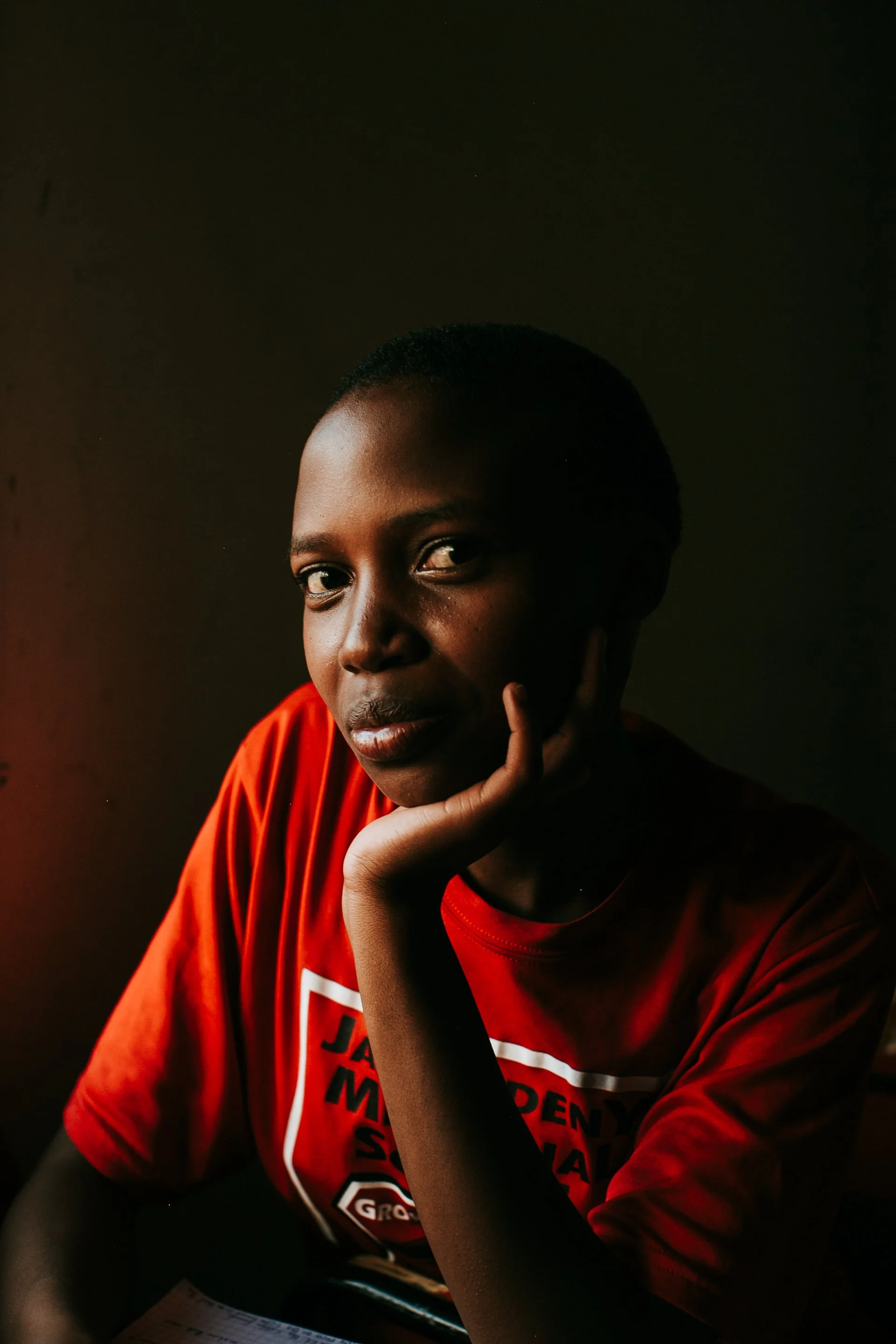 A young woman with dark skin and short hair, wearing a red T-shirt, resting her face on her left hand, looking directly at the camera with a serious expression.