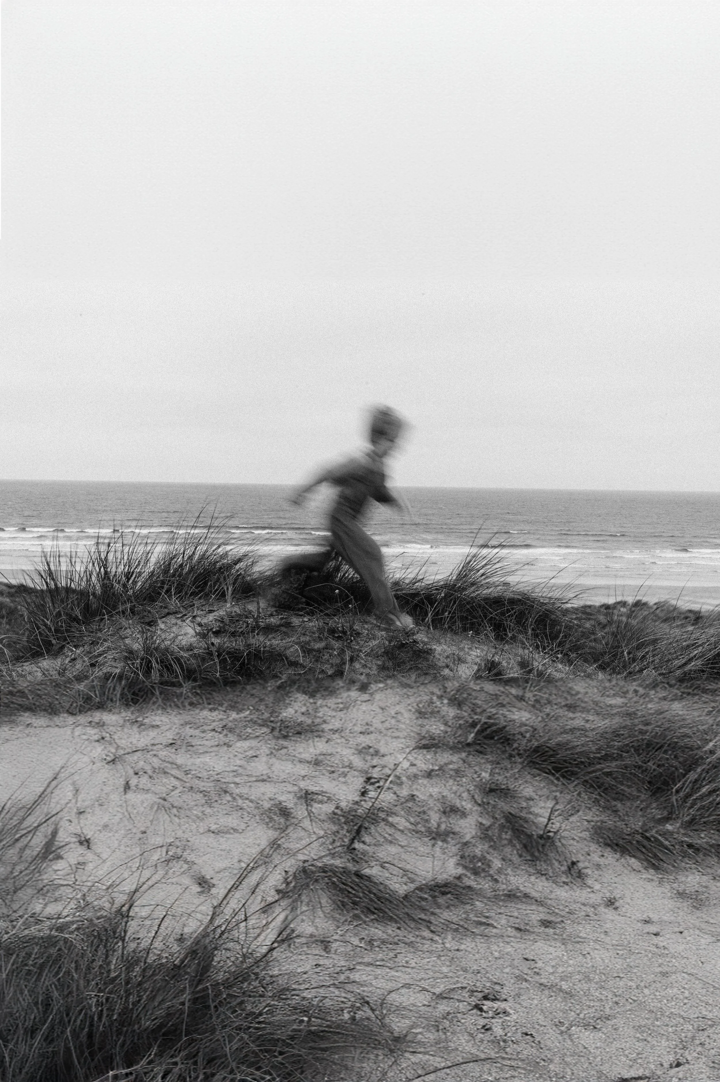 A young boy running on the sand dunes near the beach with ocean waves in the background, in black and white.
