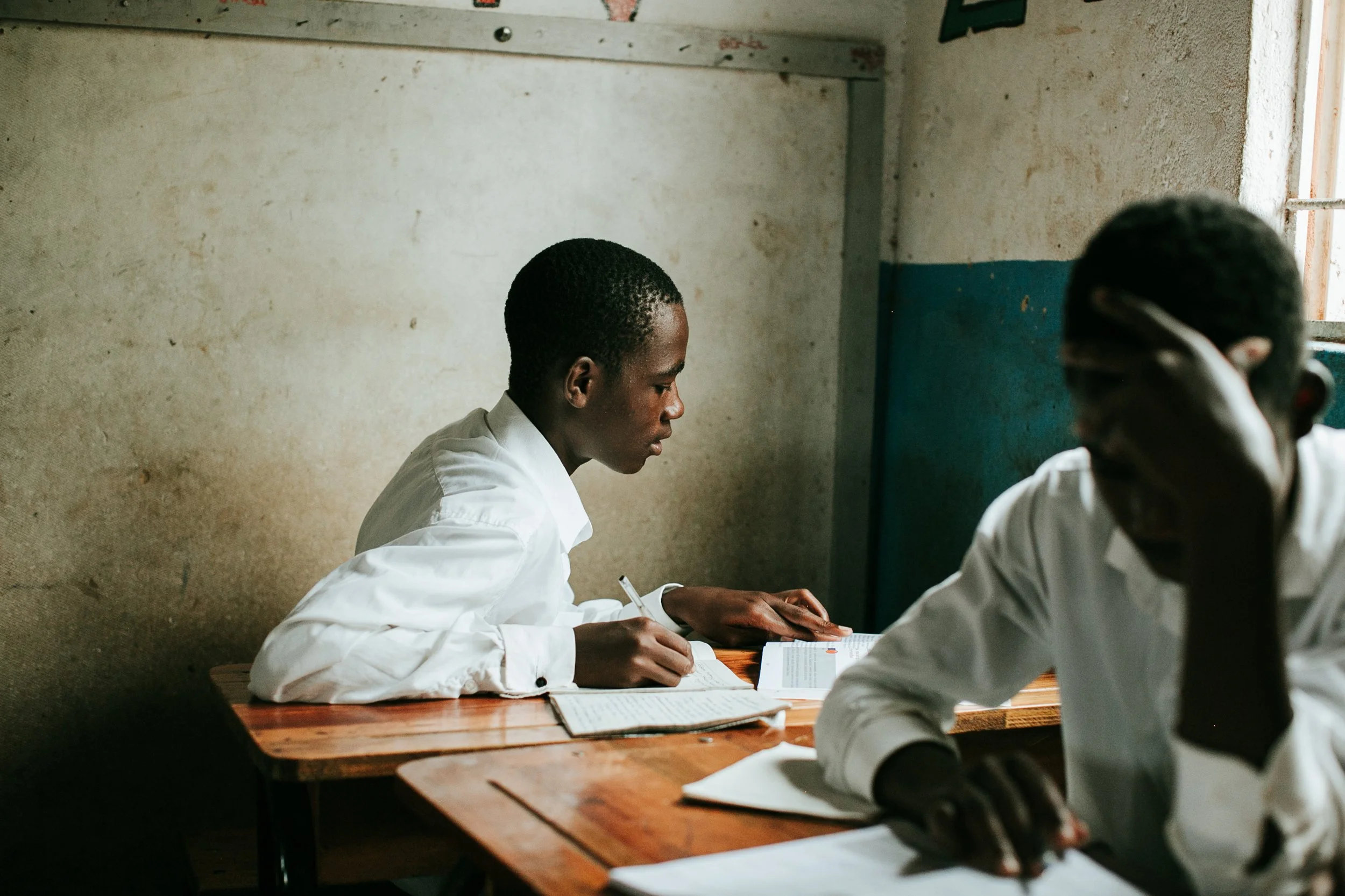 Two students in white shirts sitting at wooden desks in a classroom, one focused on writing and the other resting with his hand on his face.