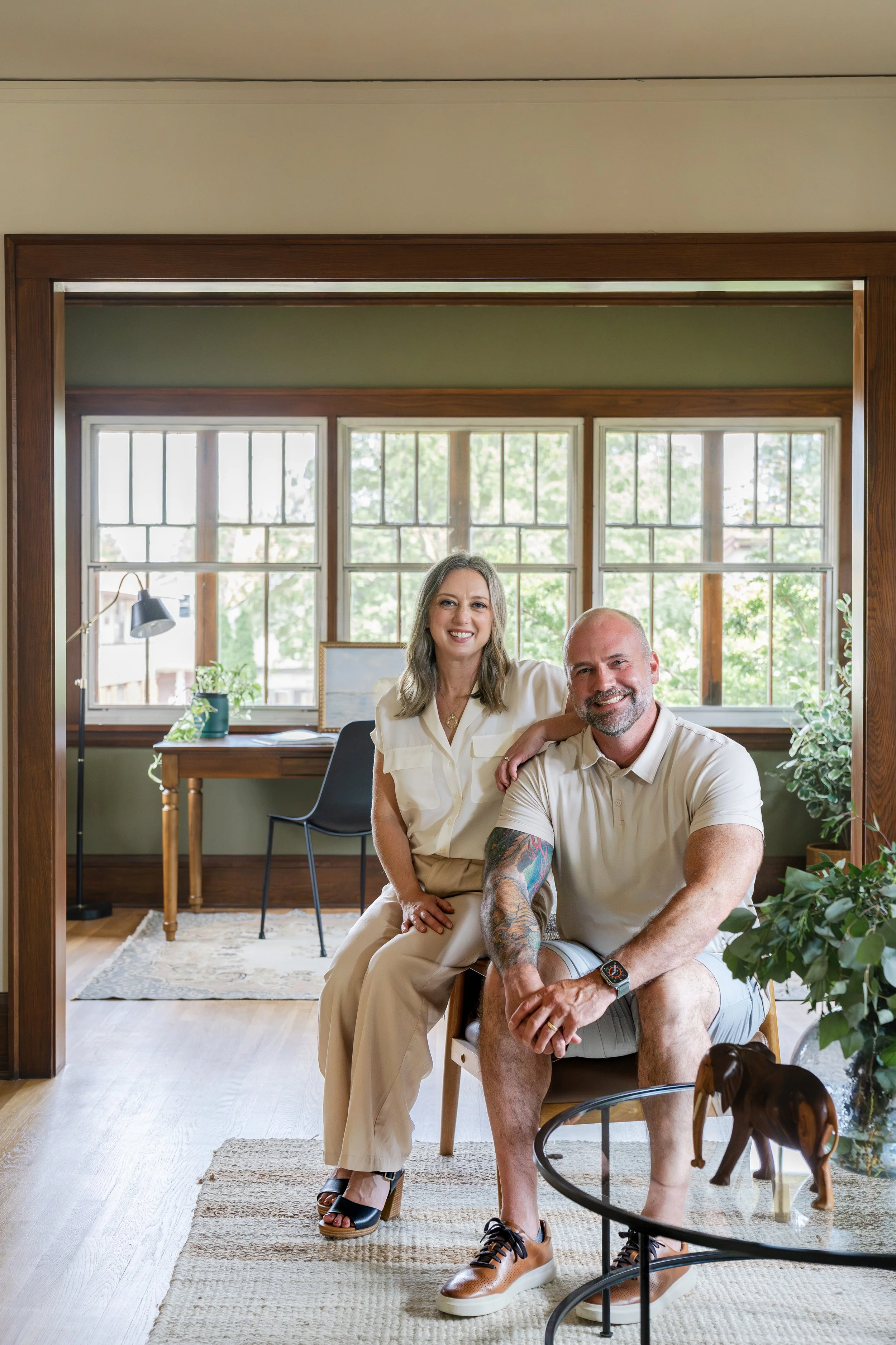 A smiling couple sits in a sunlit room with large windows and green walls. The woman has shoulder-length hair, wears a cream blouse and beige pants, and rests her hand on the man's shoulder. The man has a beard, tattoos, and wears a light-colored pol