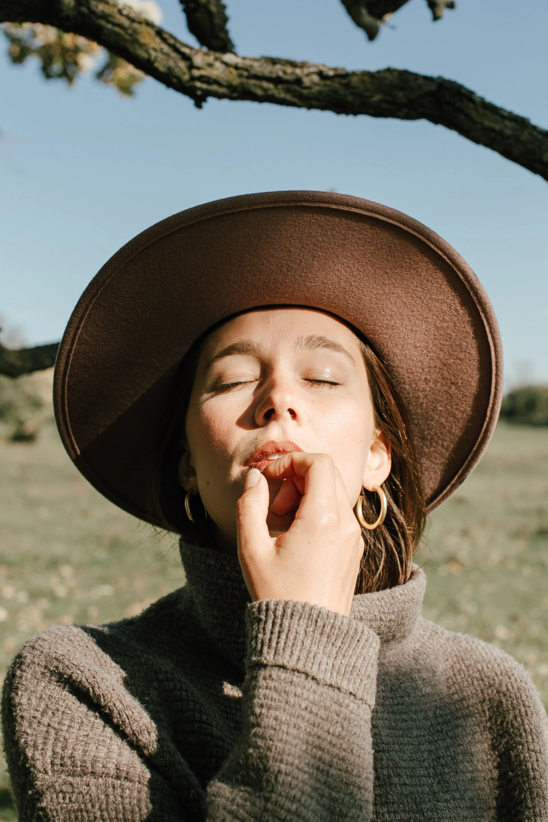 A woman in a beige sweater and large brown hat with her eyes closed, outdoors with a tree branch above her, and sunlight illuminating her face.
