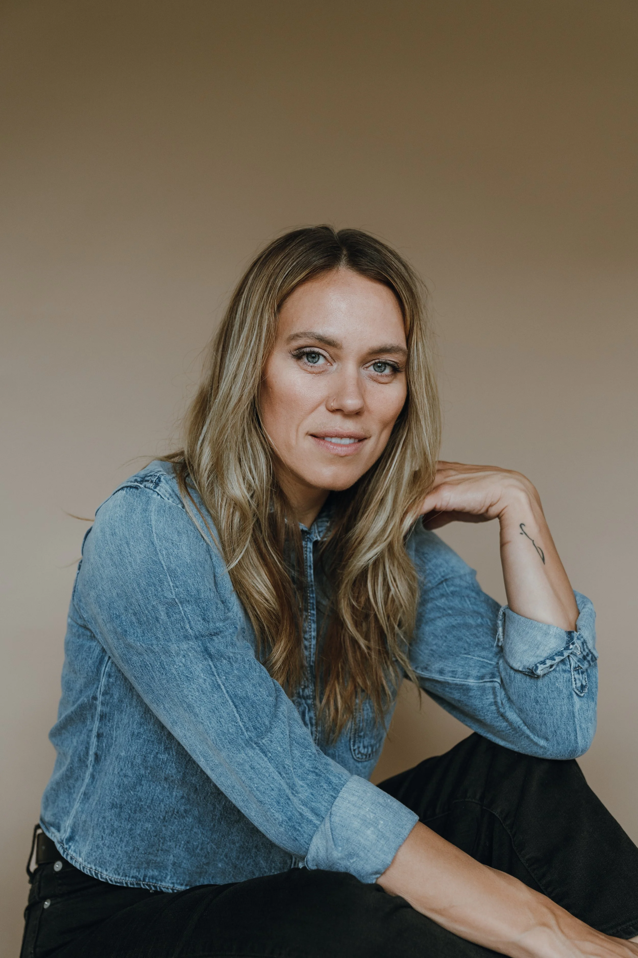 A woman with long blonde hair, blue eyes, and a nose piercing, wearing a denim jacket and black pants, sitting against a plain beige background.