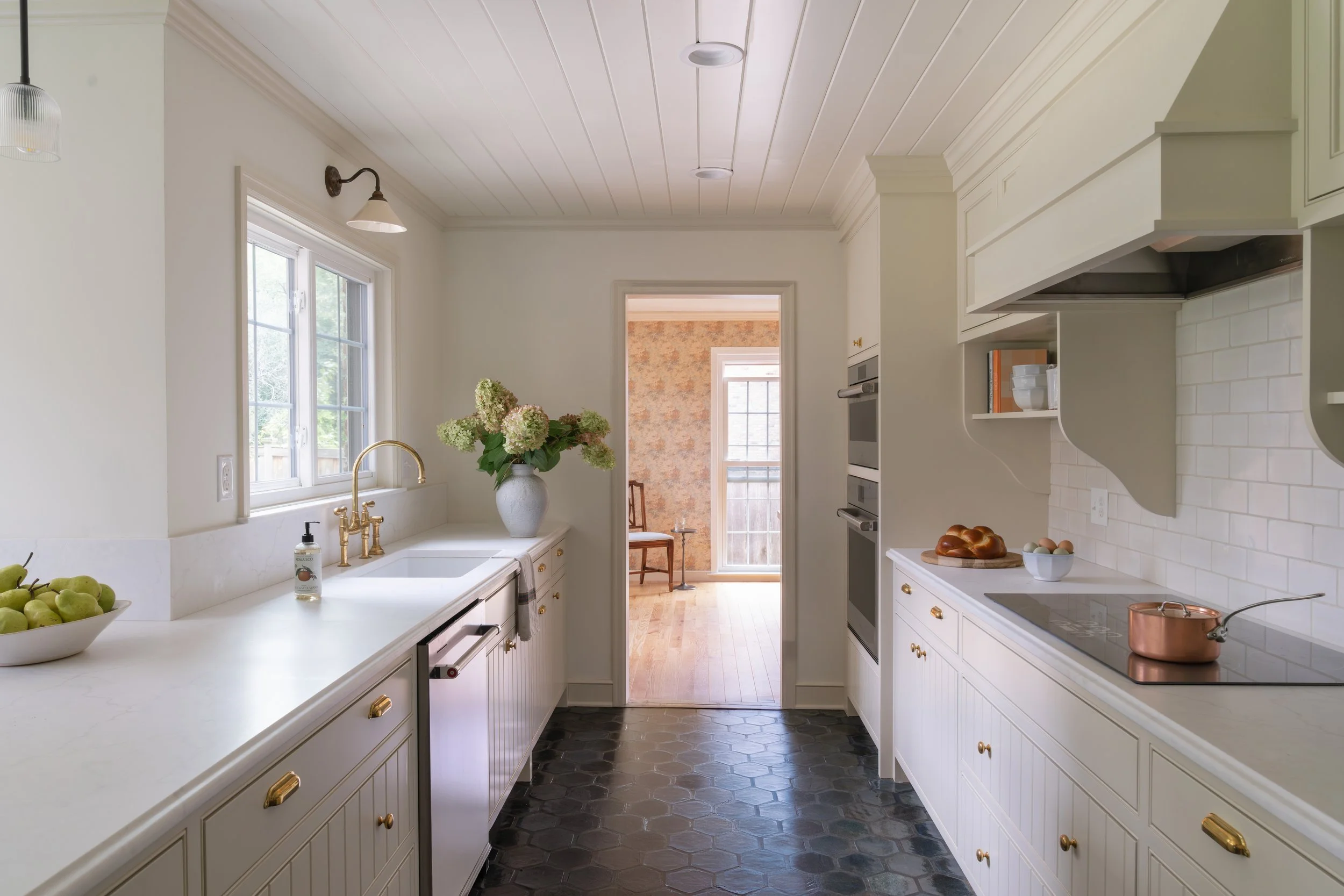 Bright kitchen with white cabinets, black hexagon tile floor, gold hardware, and a window above the sink with a vase of flowers and green apples.