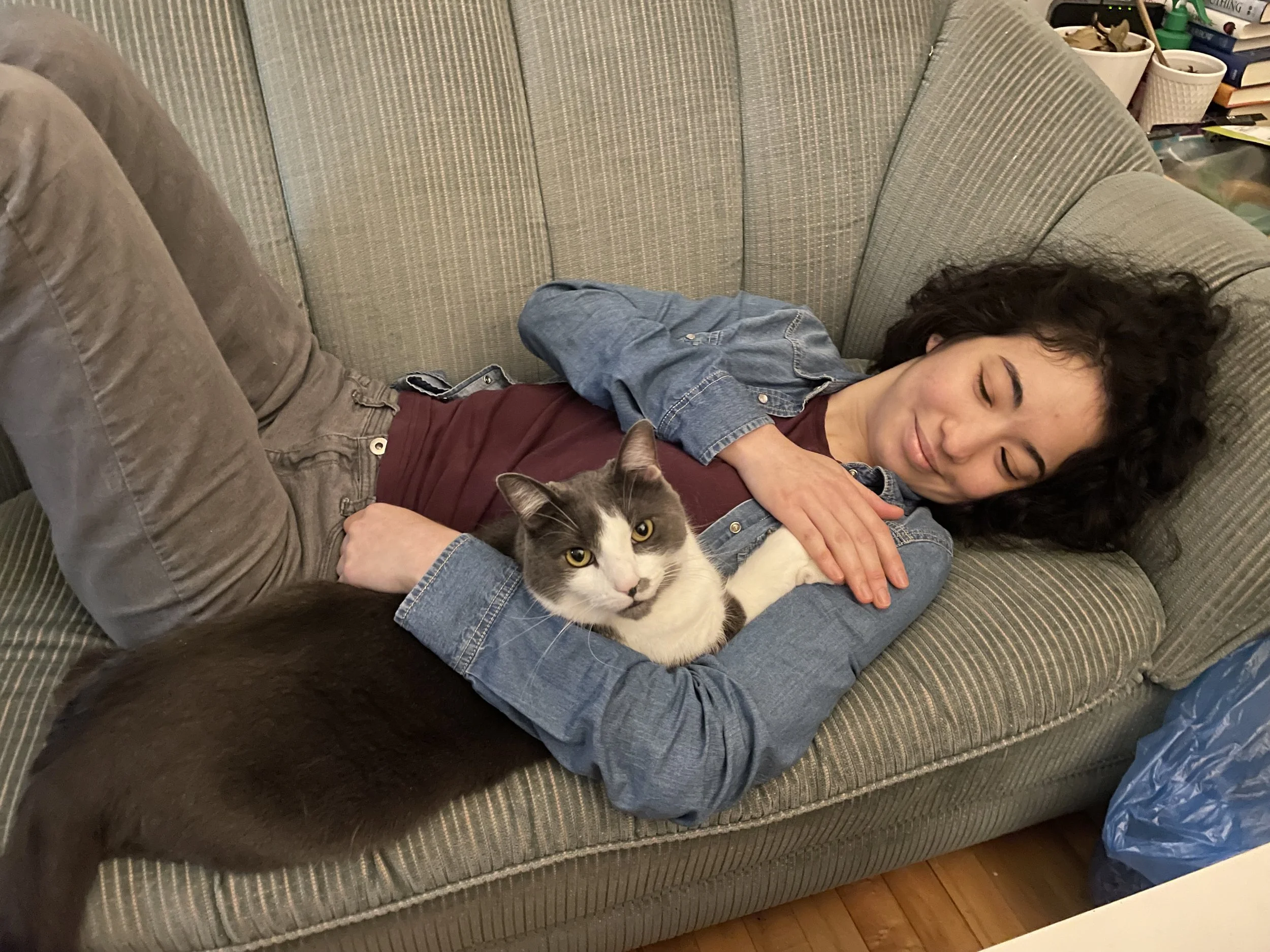 A dark-haired person lying on a green couch with a grey and white cat, both relaxing with her eyes closed.